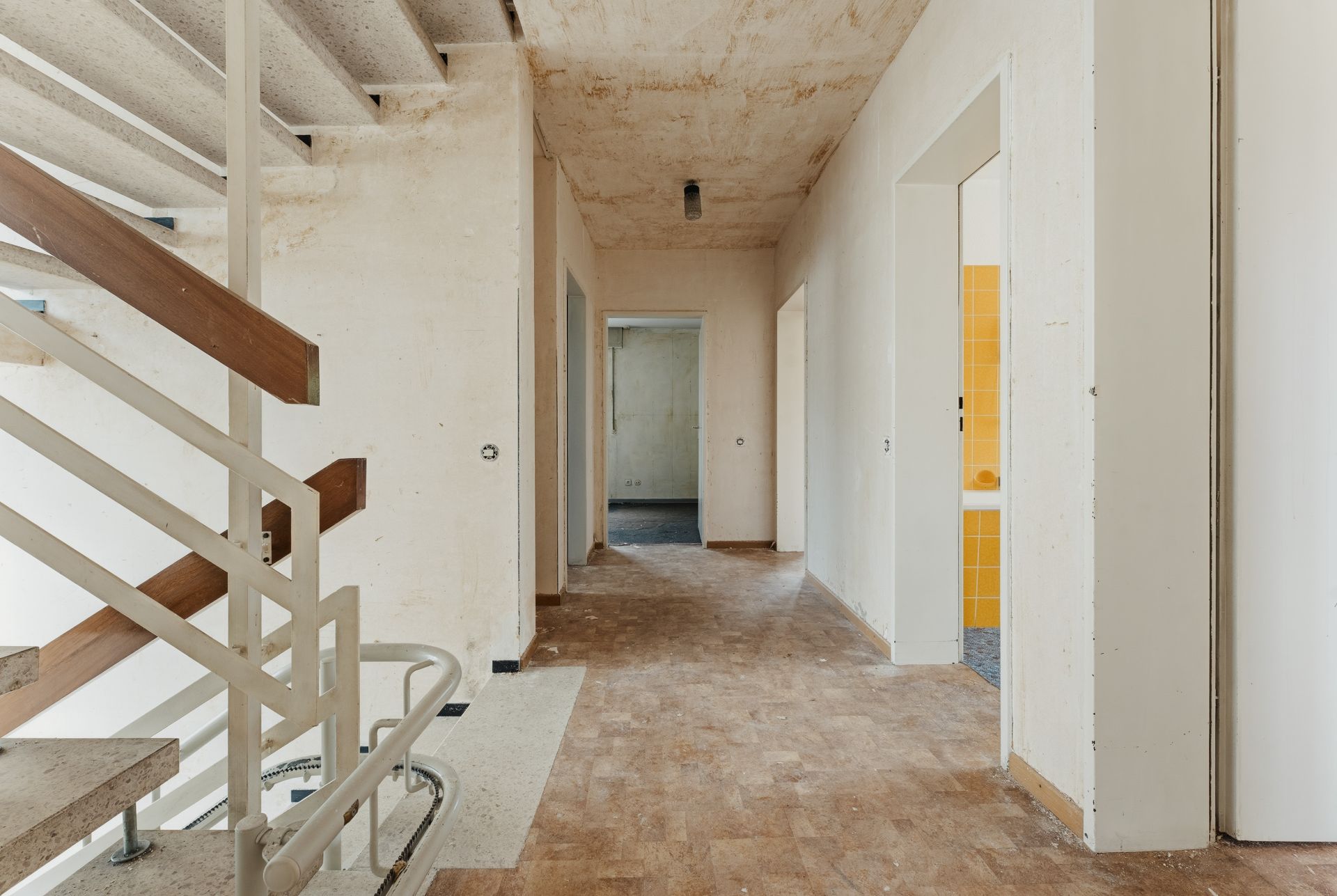 Ein leerer Flur mit offener Treppe und mehreren offenen Türen, eine Wand mit gelben Fliesen sichtbar.