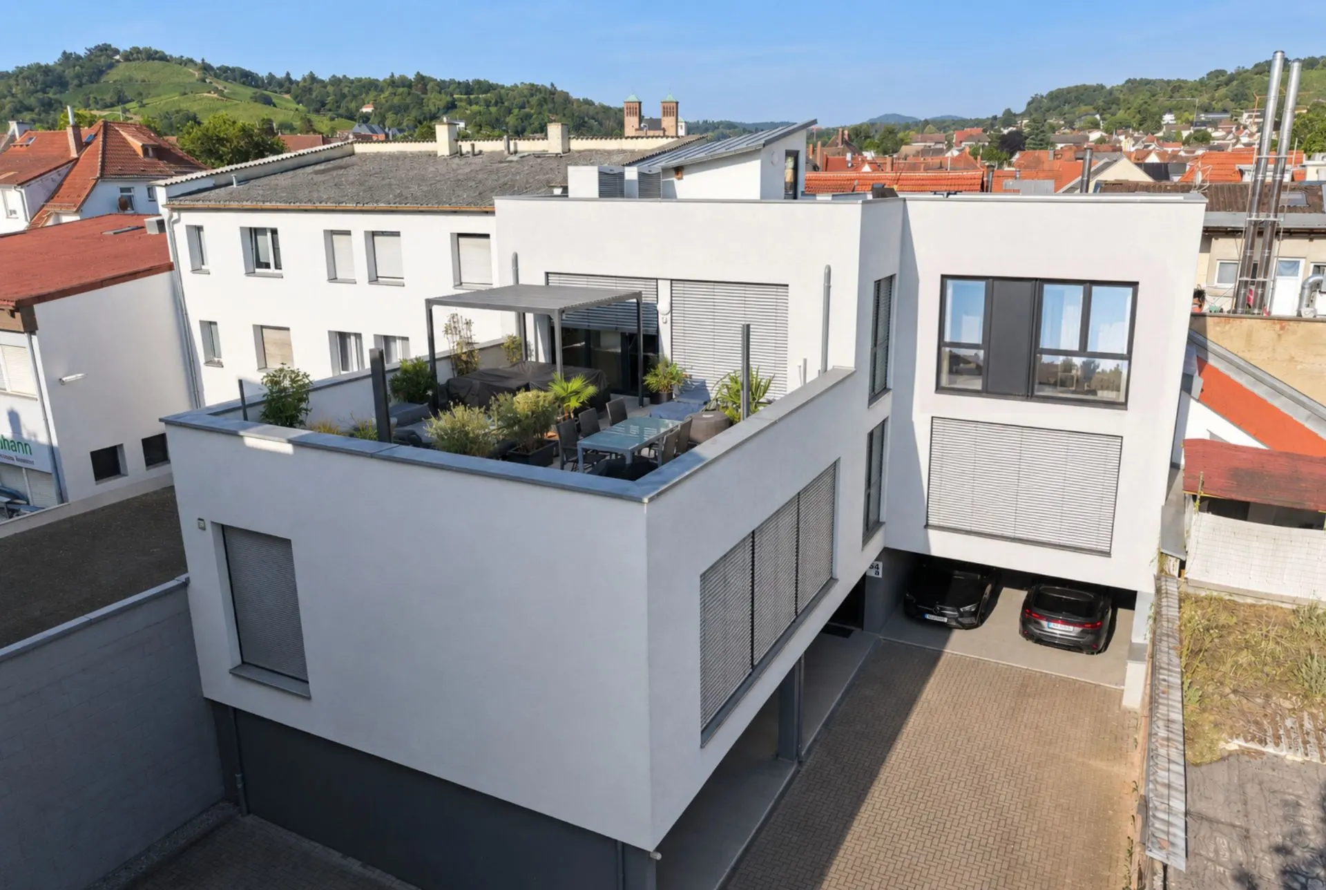 Modernes Wohnhaus mit großer Dachterrasse und darunterliegendem Carport, umgeben von weiteren Gebäuden.