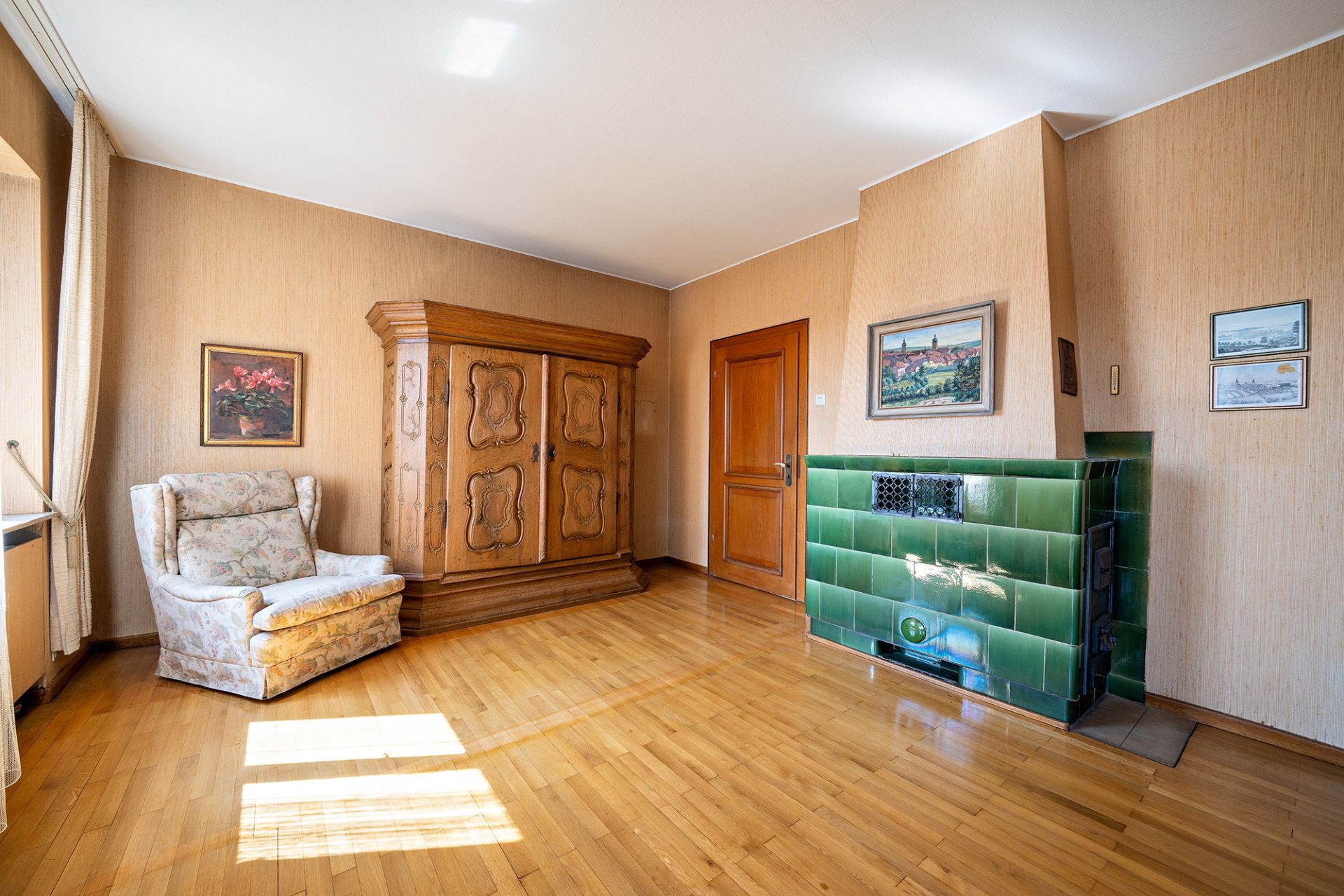 Helles Wohnzimmer mit Sessel, Holzschrank und grünem Kachelofen auf Parkettboden.