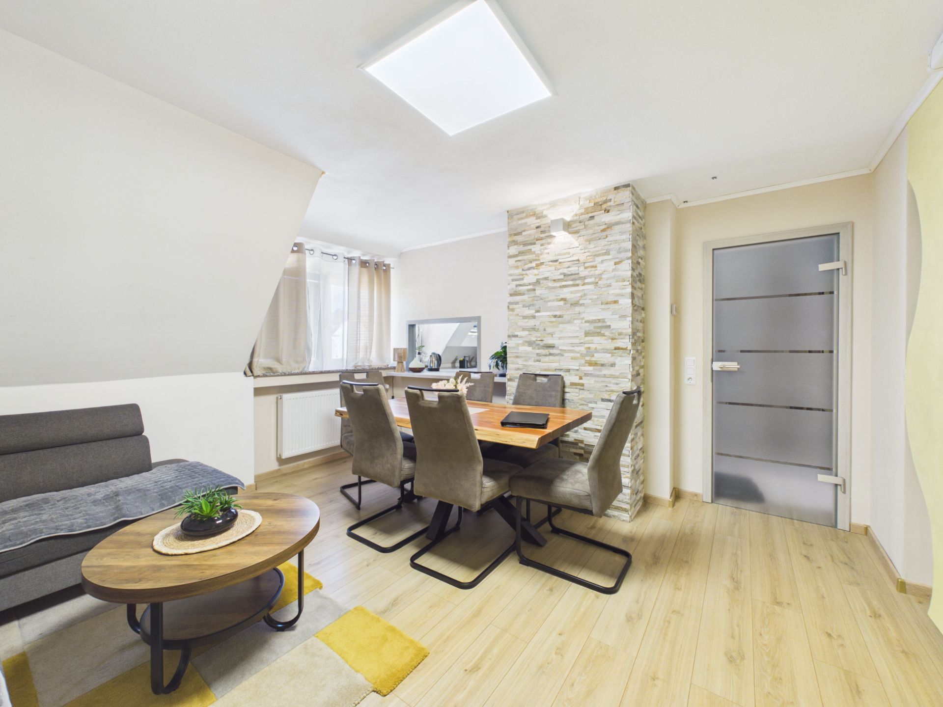 Helles Wohnzimmer mit grauem Sofa, Holztisch und Essbereich mit sechs Stühlen.
