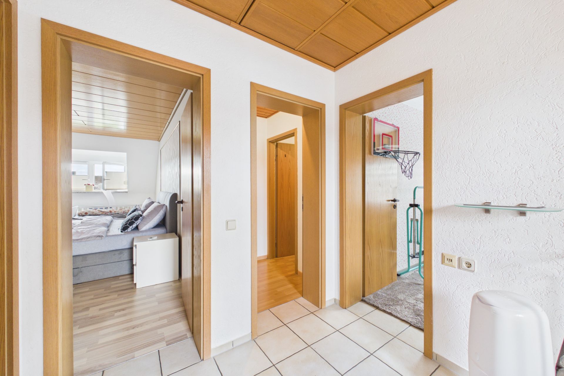 Heller Flur mit drei offenen Holztüren zu verschiedenen Zimmern, eines mit Bett, eines mit Basketballkorb.