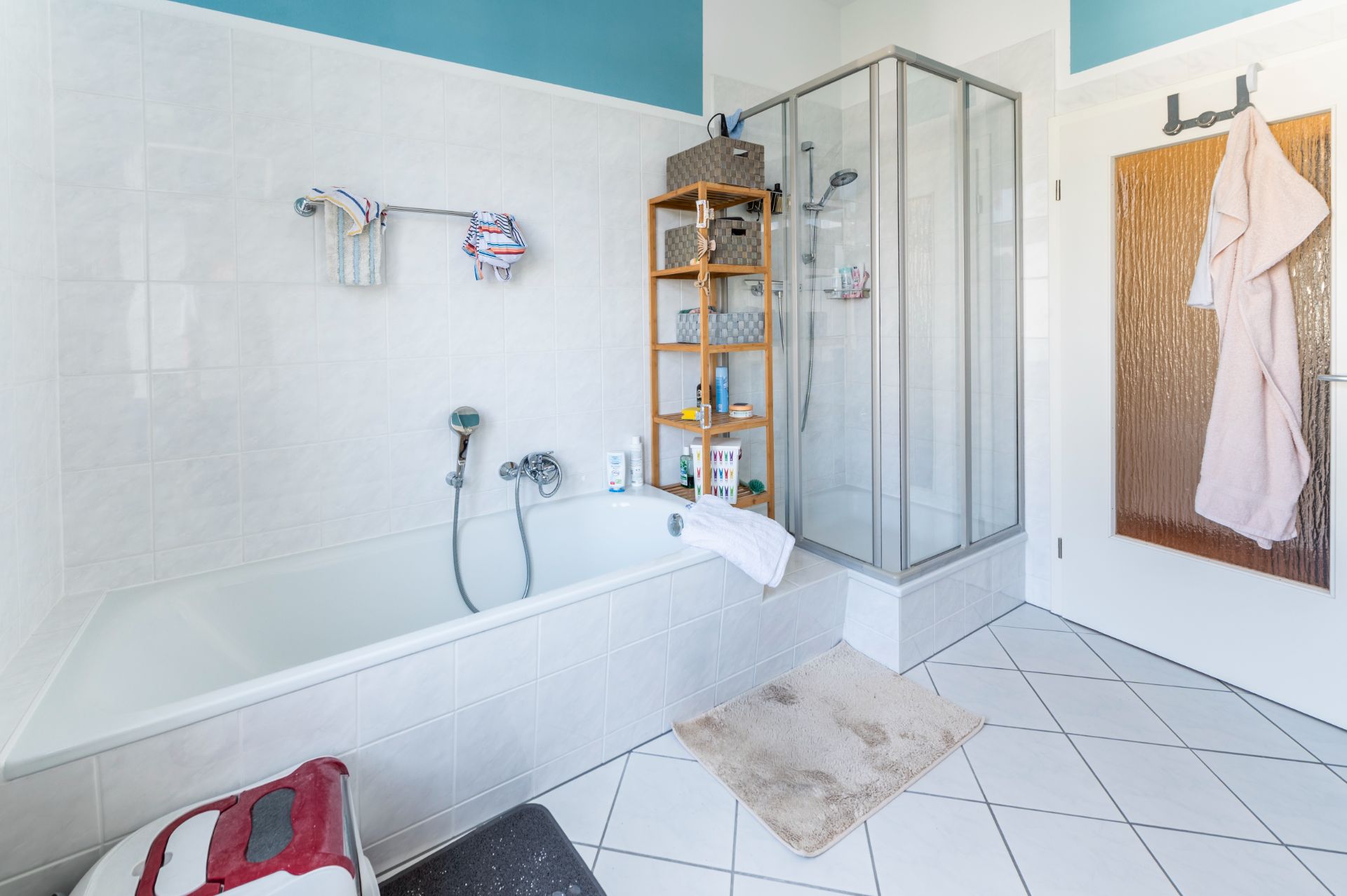 Helles Badezimmer mit Badewanne, Dusche, Regal, Handtuch und Bademantel an der Tür.