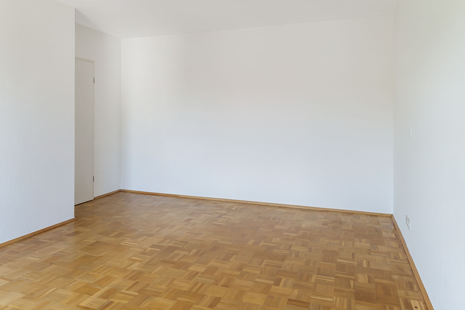 Leerer Raum mit Parkettboden und weißen Wänden, Tür links im Bild.