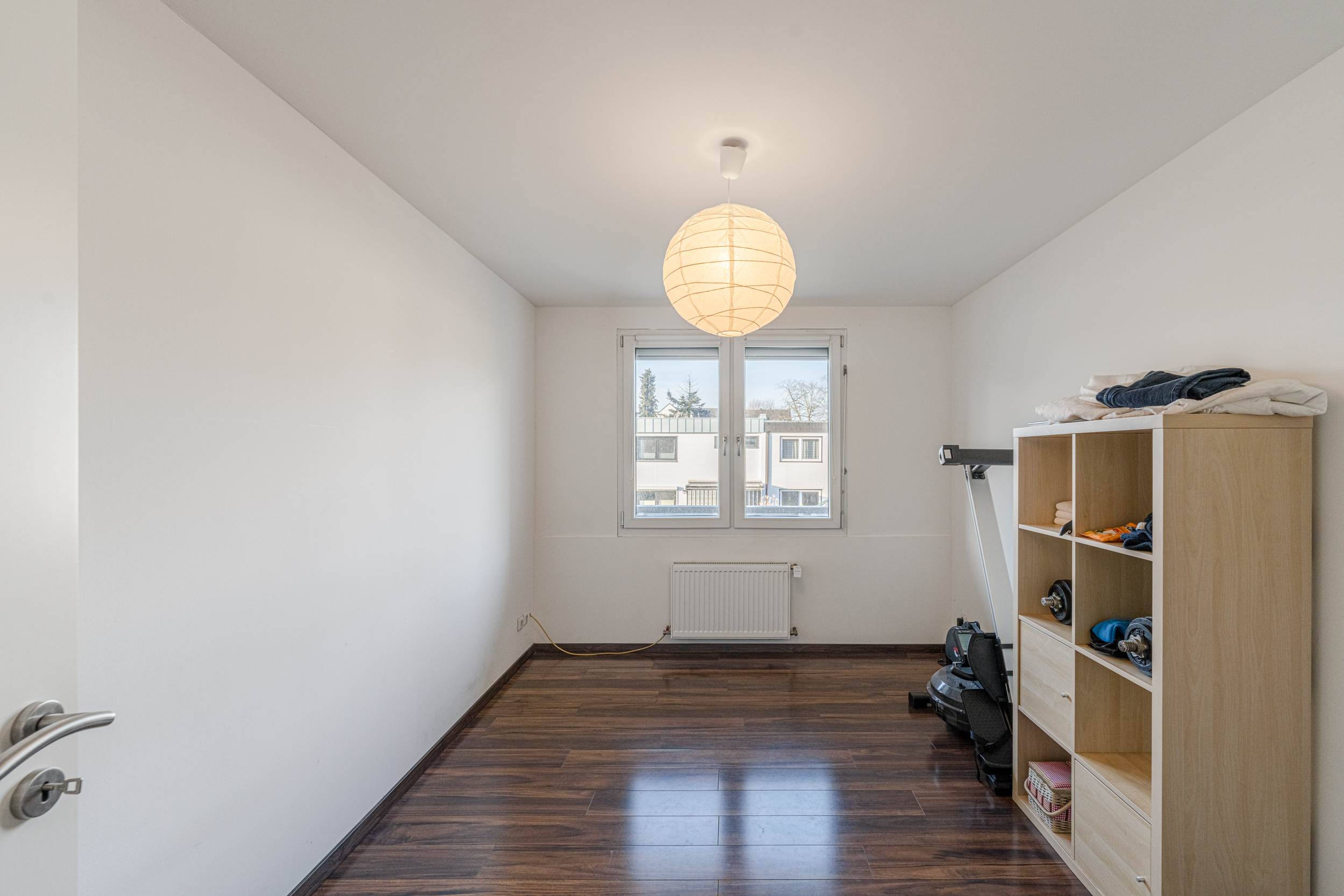Helles Zimmer mit Holzfußboden, Regal, Fenster und Deckenlampe.