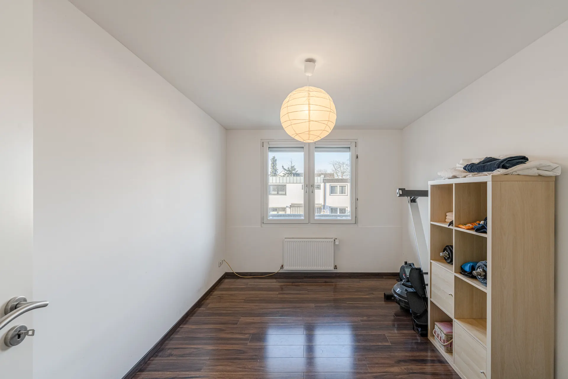 Helles Zimmer mit Holzfußboden, Regal, Fenster und Deckenlampe.
