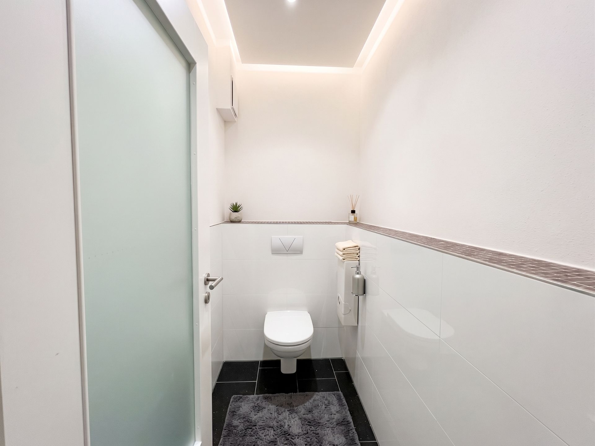 Modernes, weißes WC mit schwarzem Fliesenboden und grauem Teppich, Deko auf dem Sims.