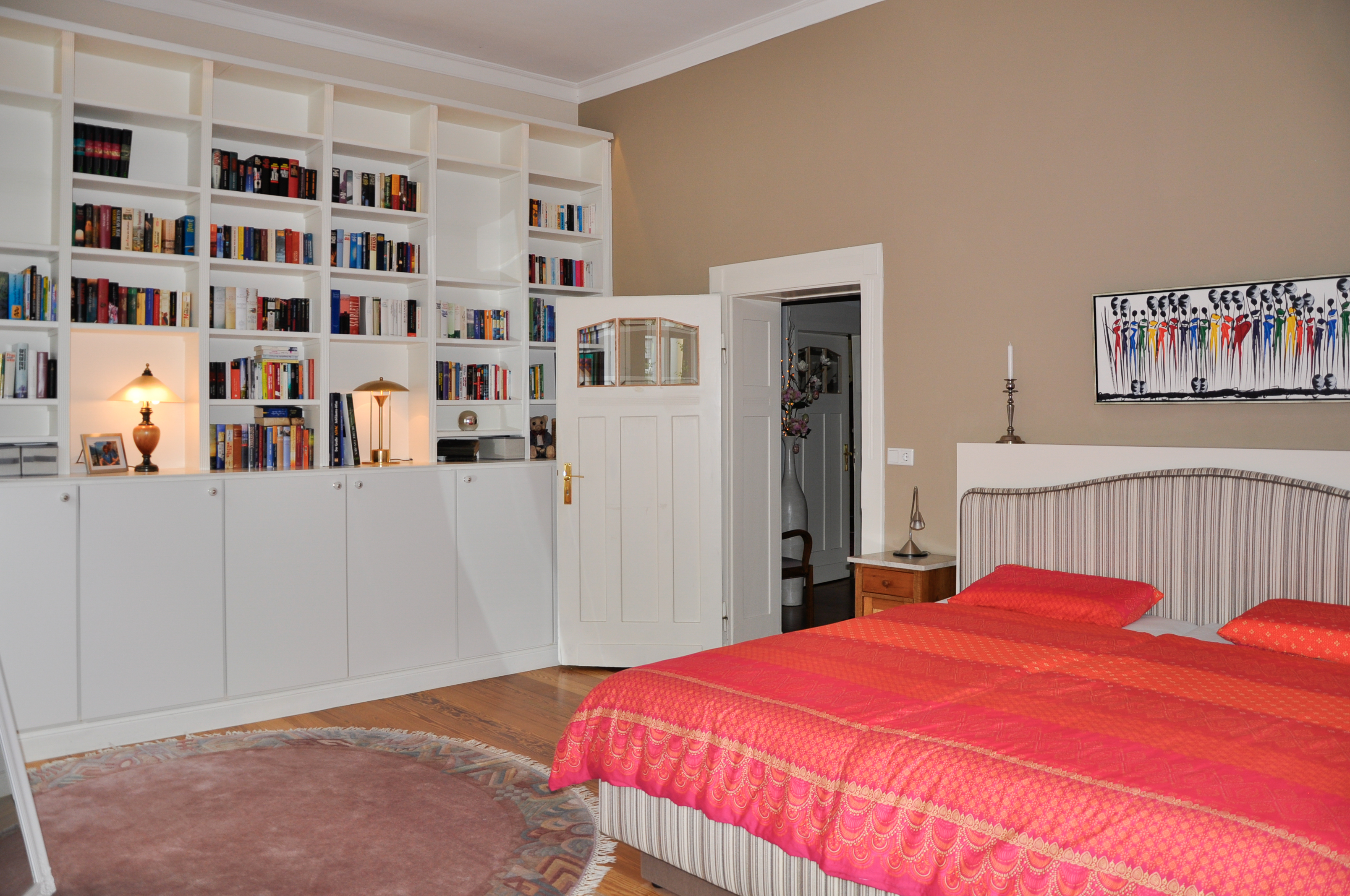 Schlafzimmer mit großem Bücherregal, Doppelbett mit roter Bettdecke und offener Tür.