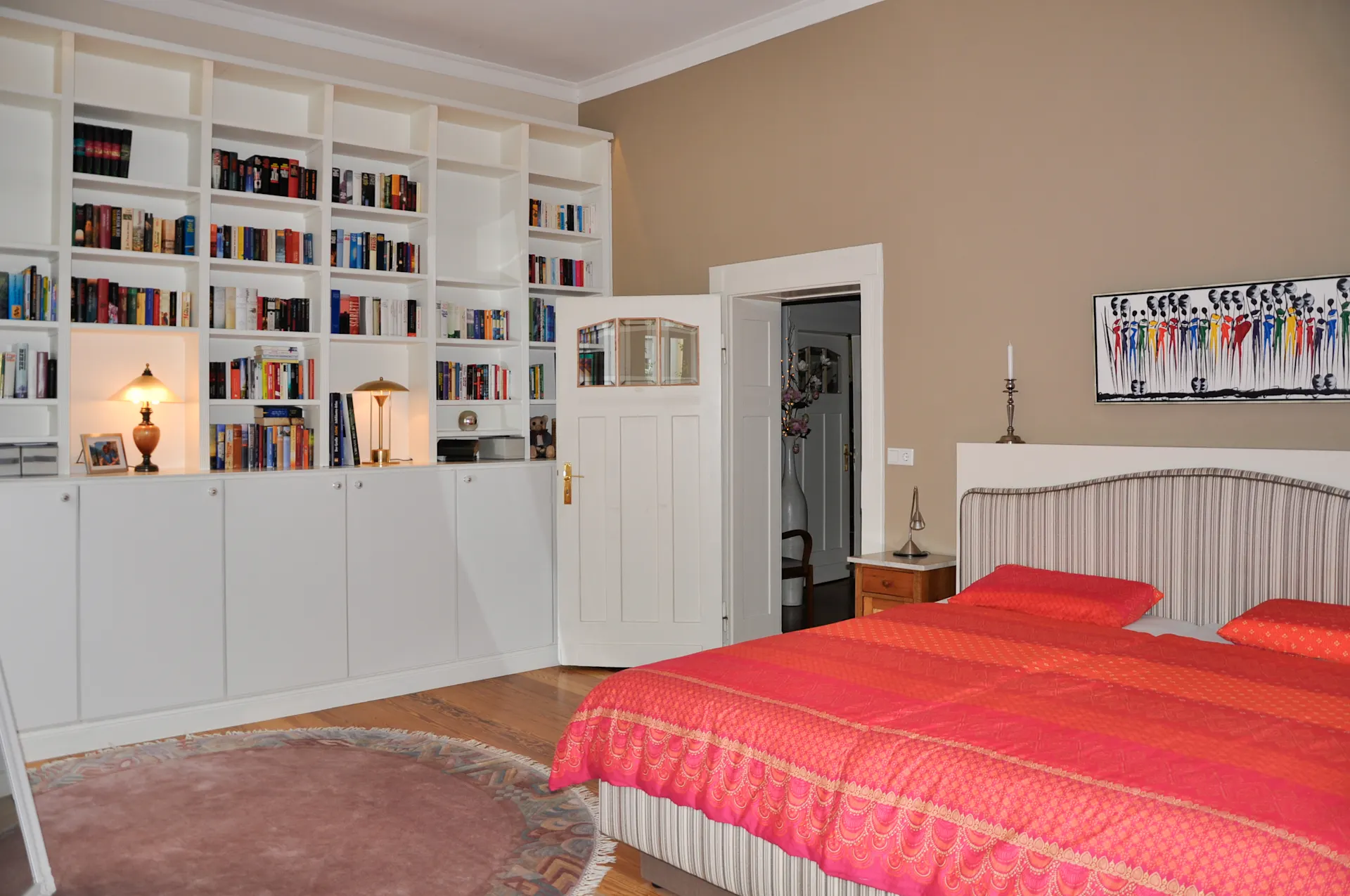 Schlafzimmer mit großem Bücherregal, Doppelbett mit roter Bettdecke und offener Tür.