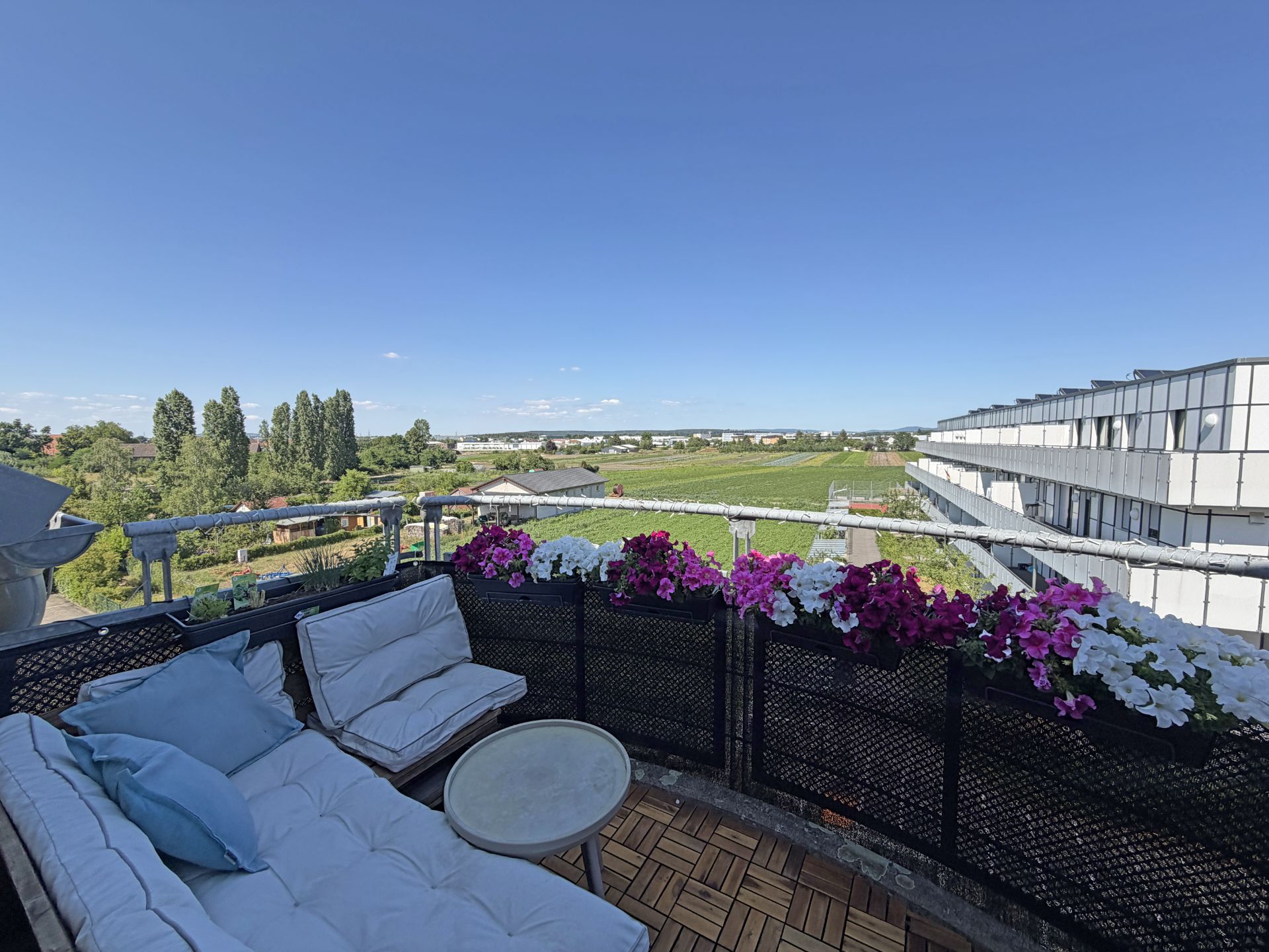 Balkon mit Sitzbank, kleinem Tisch und Blumenkästen, Blick auf grüne Landschaft und moderne Gebäude.
