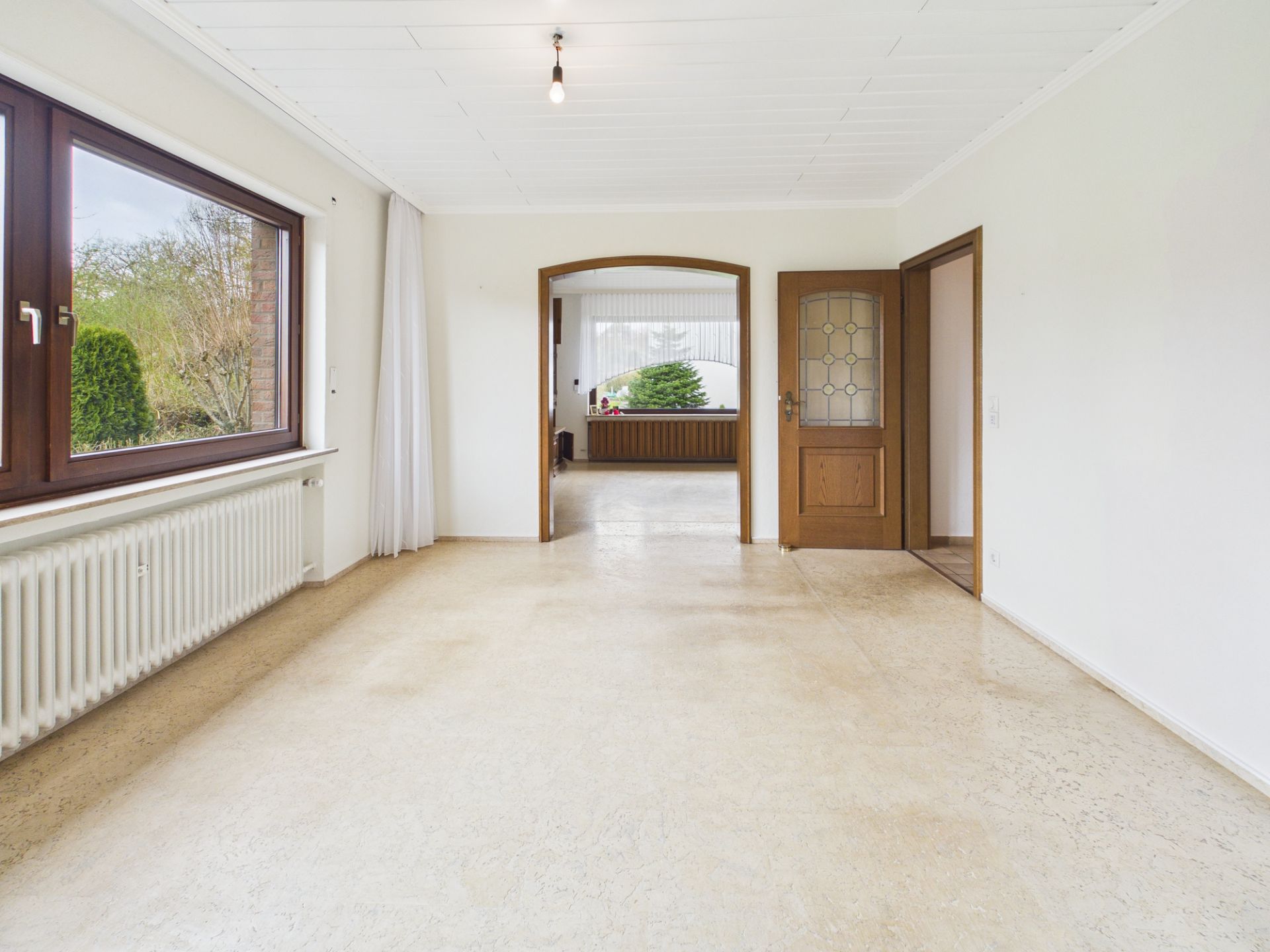 Helles, leeres Wohnzimmer mit großem Fenster, weißem Boden und Holztür zu weiterem Raum.