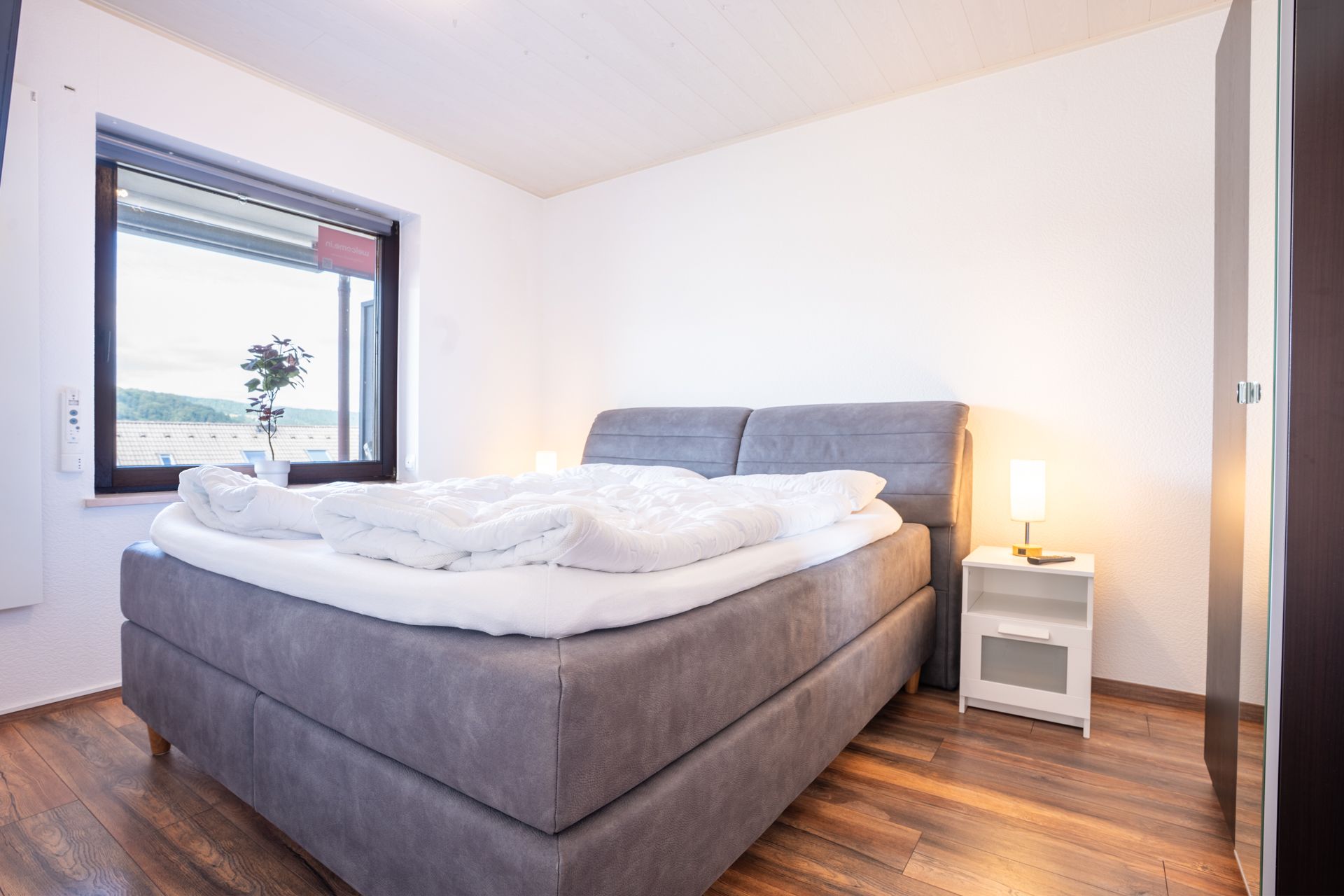 Helles Schlafzimmer mit großem grauen Bett, Nachttisch, Lampe und Fenster mit Blick nach draußen.