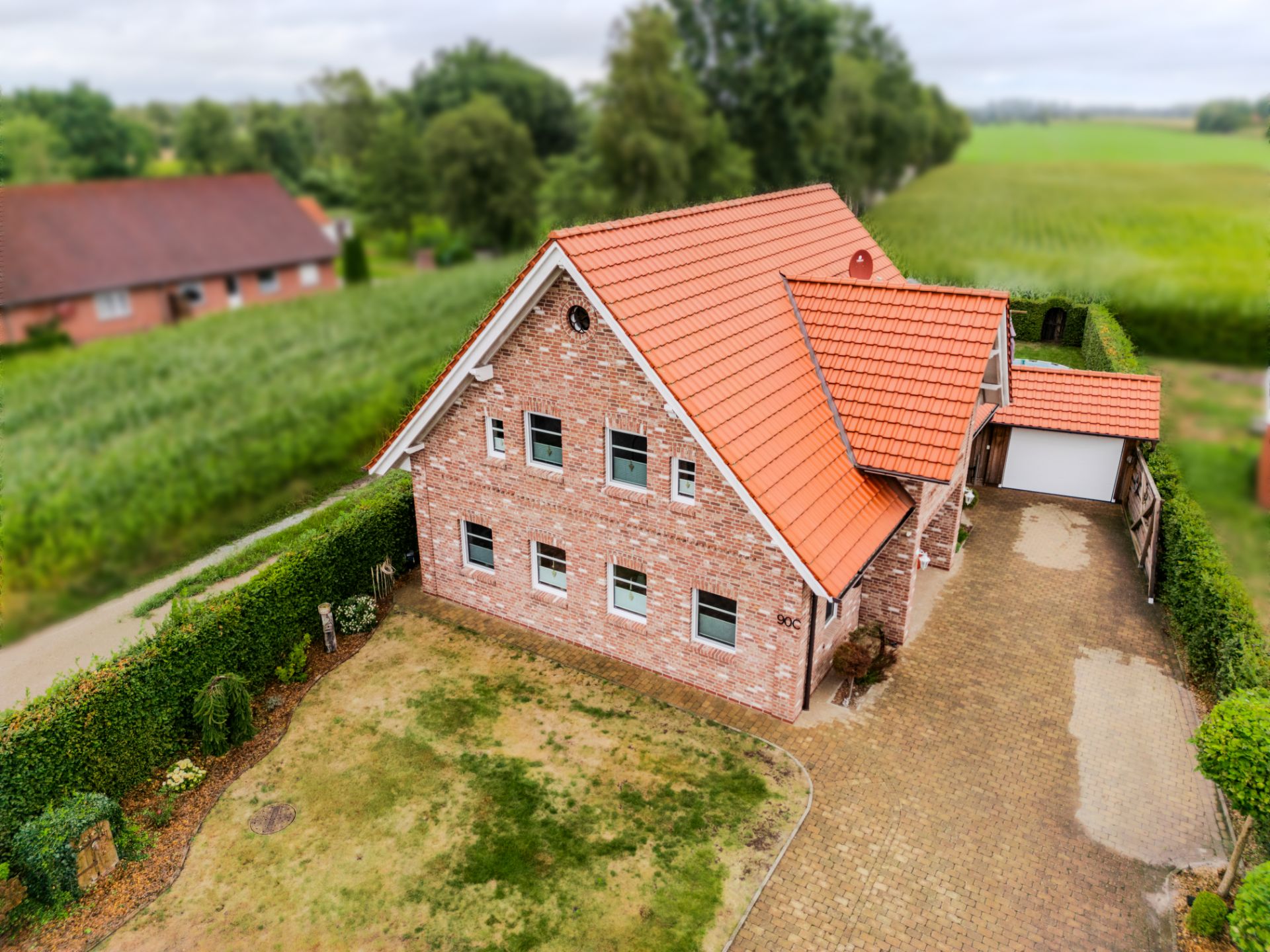 Backsteinwohnhaus mit rotem Dach, Einfahrt, kleinem Garten und umgebender Hecke.