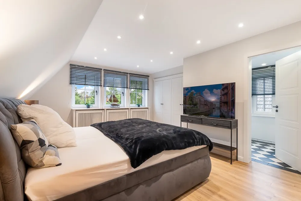 Modernes Schlafzimmer mit großem Bett, Fernseher, drei Fenstern und Blick ins angrenzende Zimmer.