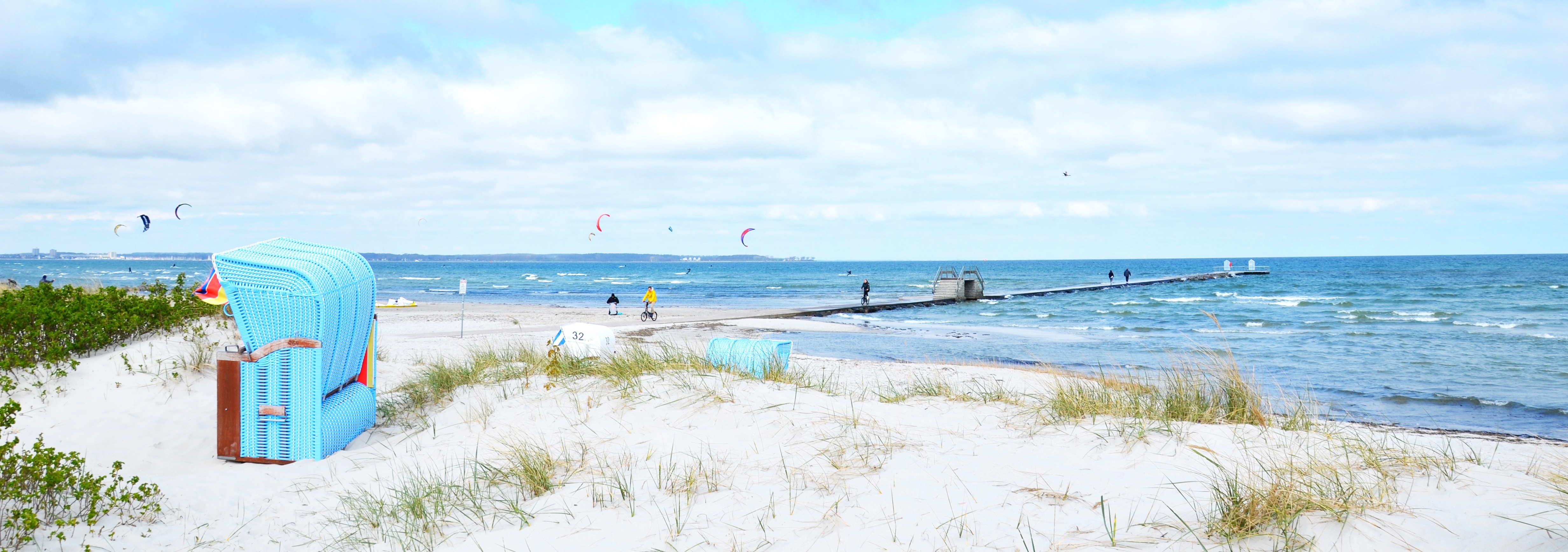 Sandstrand mit Strandkorb, Steg und Menschen im Hintergrund am Meer.