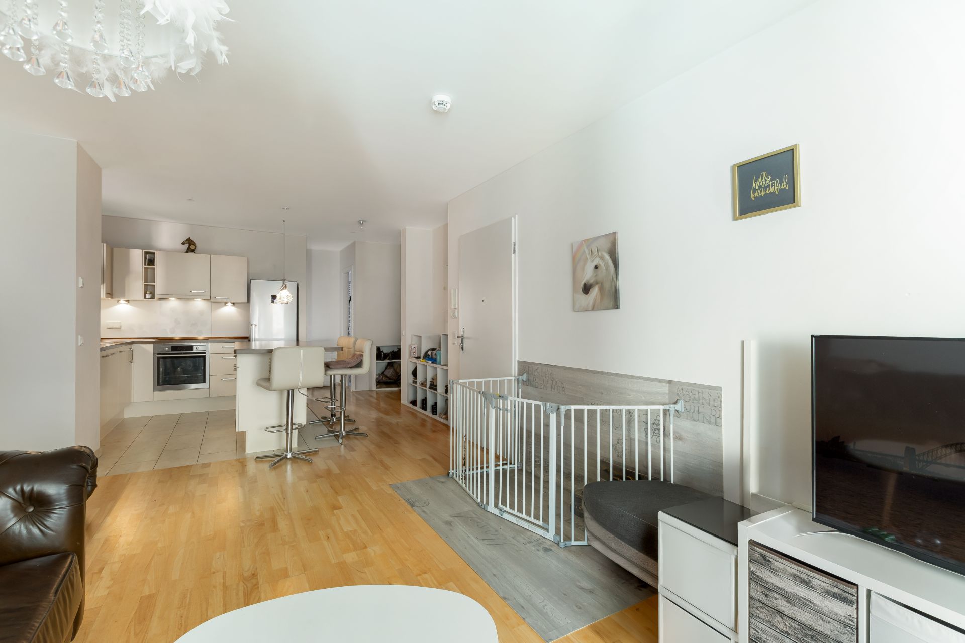 Helles Wohnzimmer mit Fernseher, Sitzbereich, Gitter, offener Küche und Holzboden.