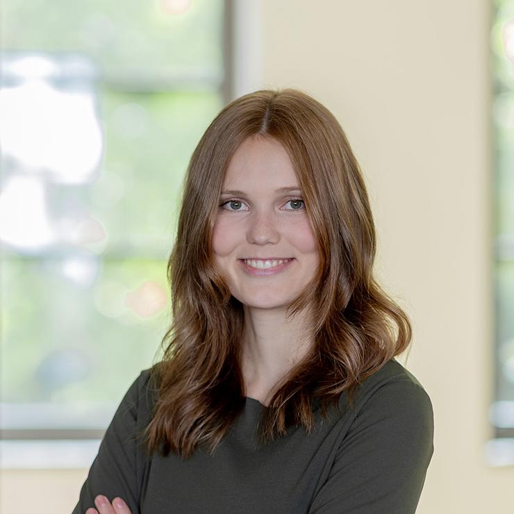 Junge Frau mit langen braunen Haaren steht mit verschränkten Armen vor einem Fenster.