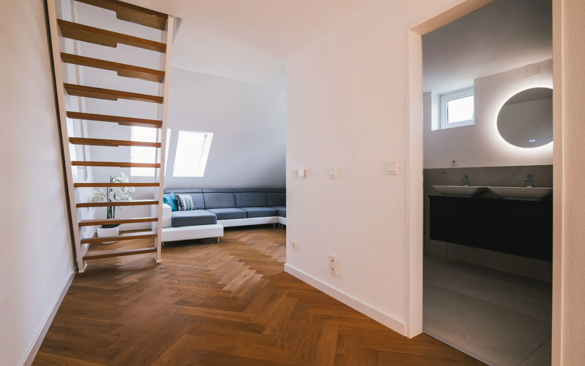 Heller Raum mit Holztreppe, grauem Sofa und Blick ins Badezimmer mit rundem Spiegel.