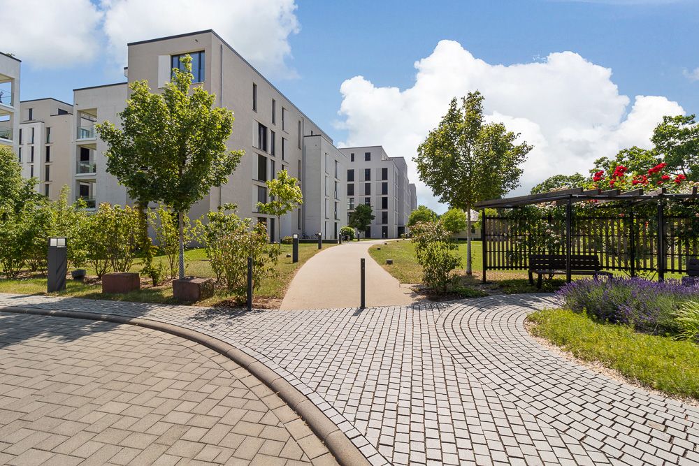 Moderne Wohngebäude mit Gehweg, Bäumen und einer Sitzbank unter einer Pergola im Grünen.