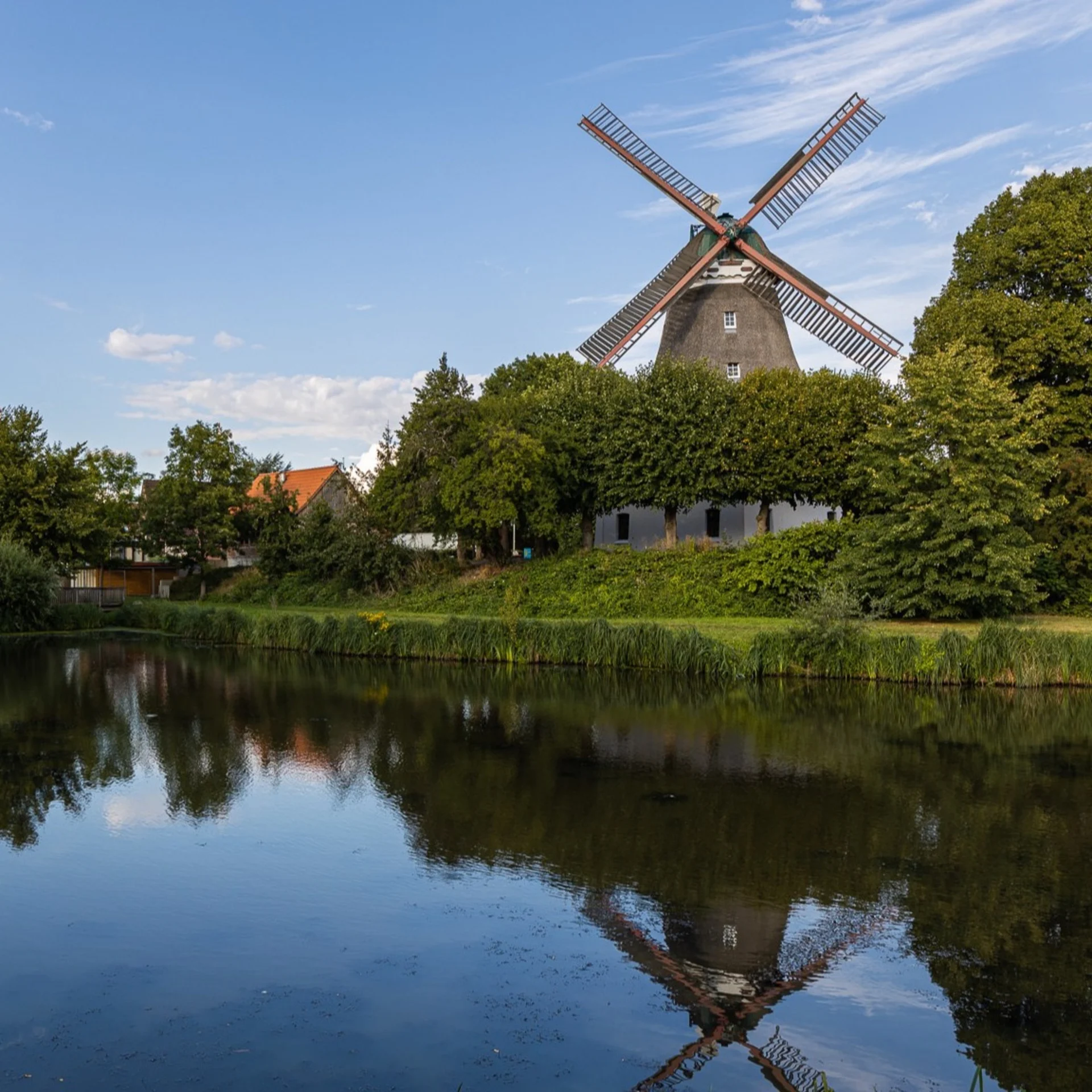 Windmühle und Wohnhäuser an einem Teich, umgeben von Bäumen und blauem Himmel.