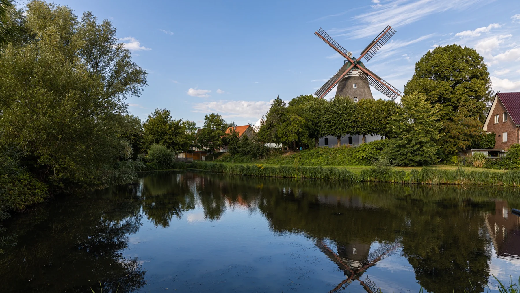 Windmühle und Wohnhäuser an einem Teich, umgeben von Bäumen und blauem Himmel.