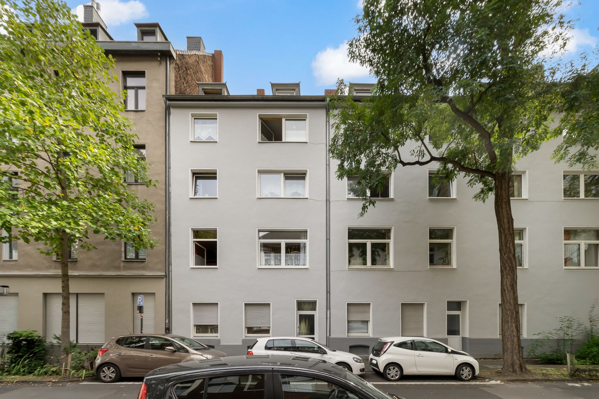 Mehrstöckiges Wohnhaus mit geparkten Autos und Bäumen an der Straße.