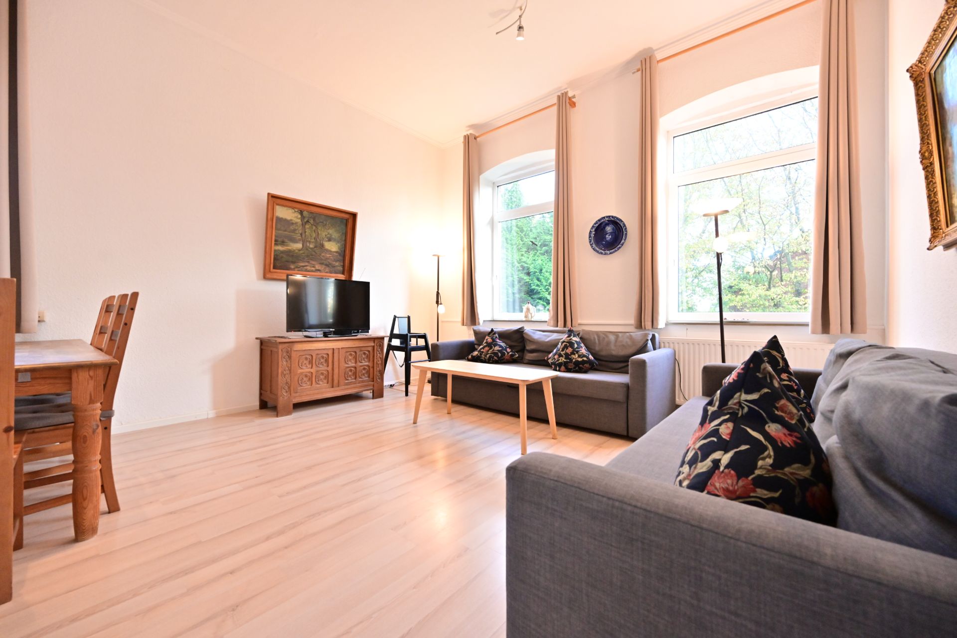 Helles Wohnzimmer mit zwei grauen Sofas, Couchtisch, Fernseher und großen Fenstern.