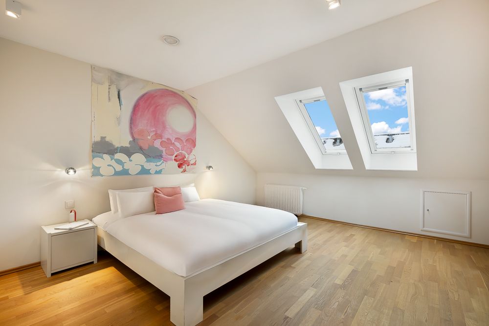 Helles Schlafzimmer mit Doppelbett, Holzfußboden, zwei Dachfenstern und Wandbild über dem Bett.