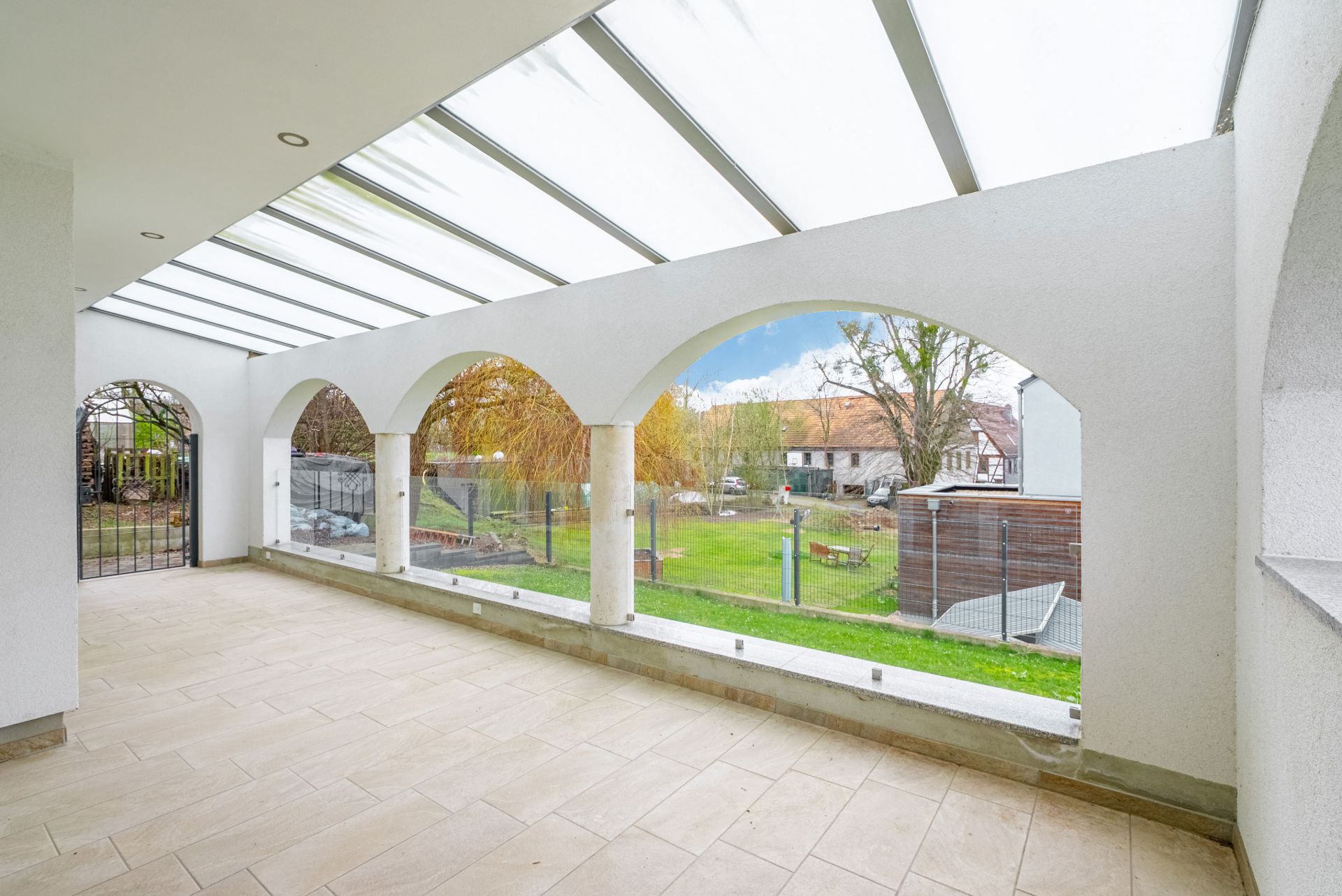 Überdachte Terrasse mit Rundbögen und Blick auf einen Garten mit Rasen und Bäumen.