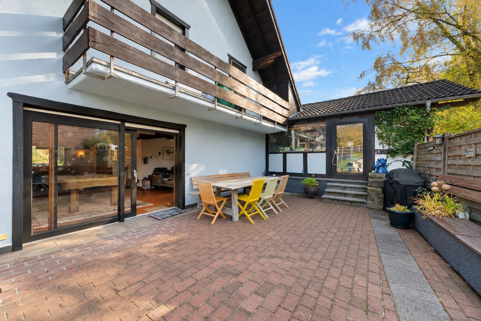 Gepflasterte Terrasse mit Holztisch, Stühlen und Zugang zu einem Haus mit Balkon.