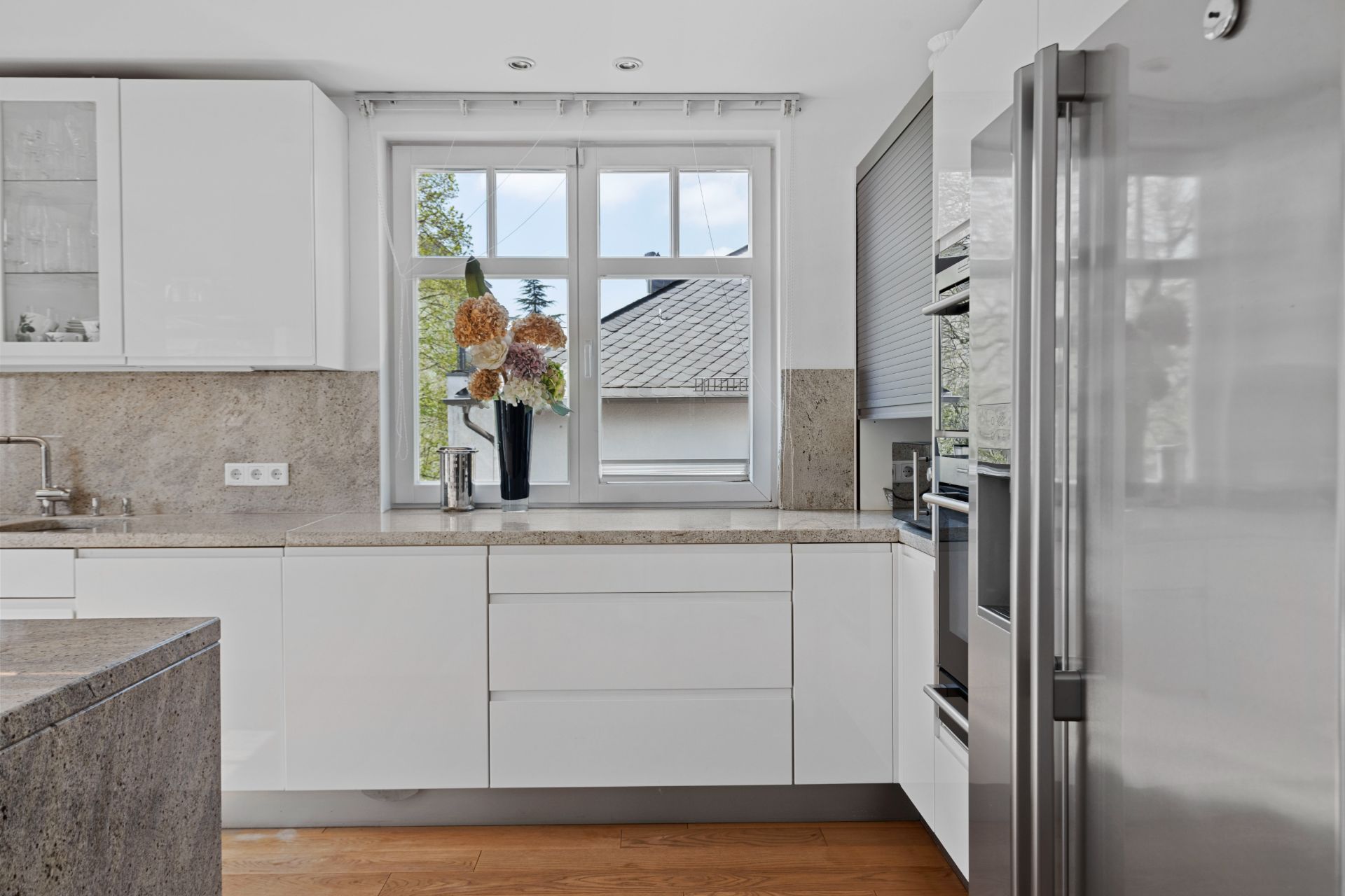 Moderne weiße Küche mit großem Fenster, Edelstahlkühlschrank und Holzfußboden.