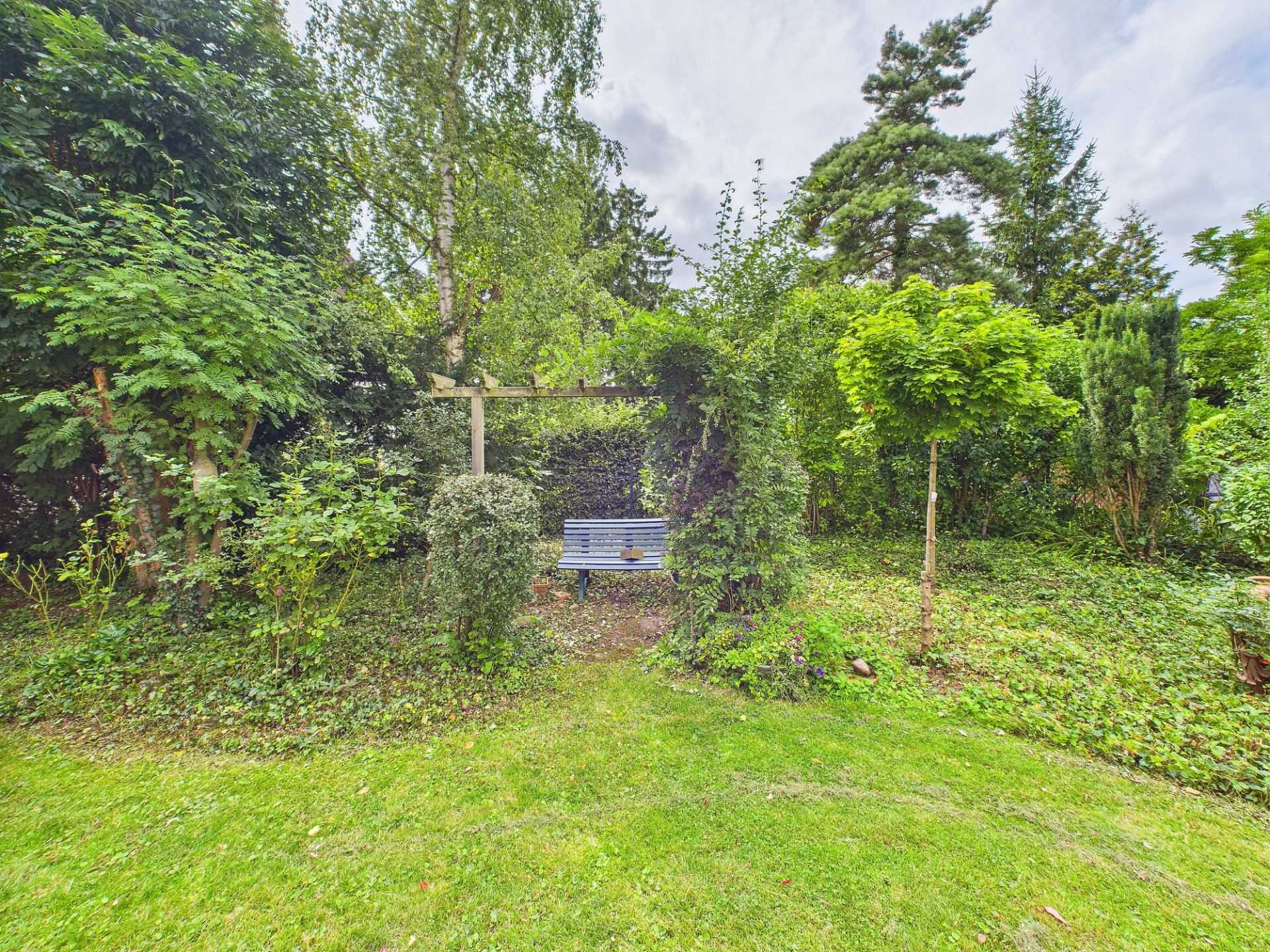 Holzbank in einem grünen Garten mit Bäumen und Sträuchern, Pergola im Hintergrund.