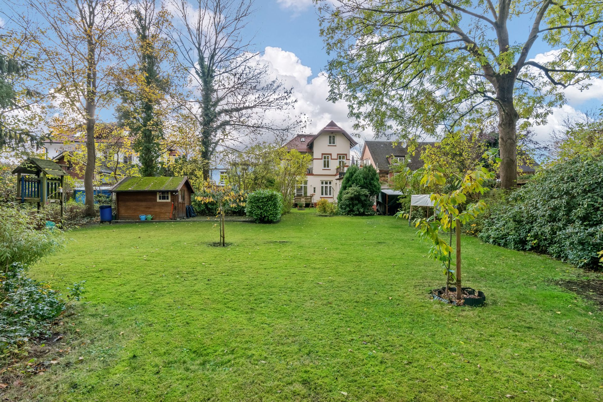 Großer Garten mit Rasen, Bäumen, Sträuchern, Spielplatz und Gartenhaus, Wohnhaus im Hintergrund.