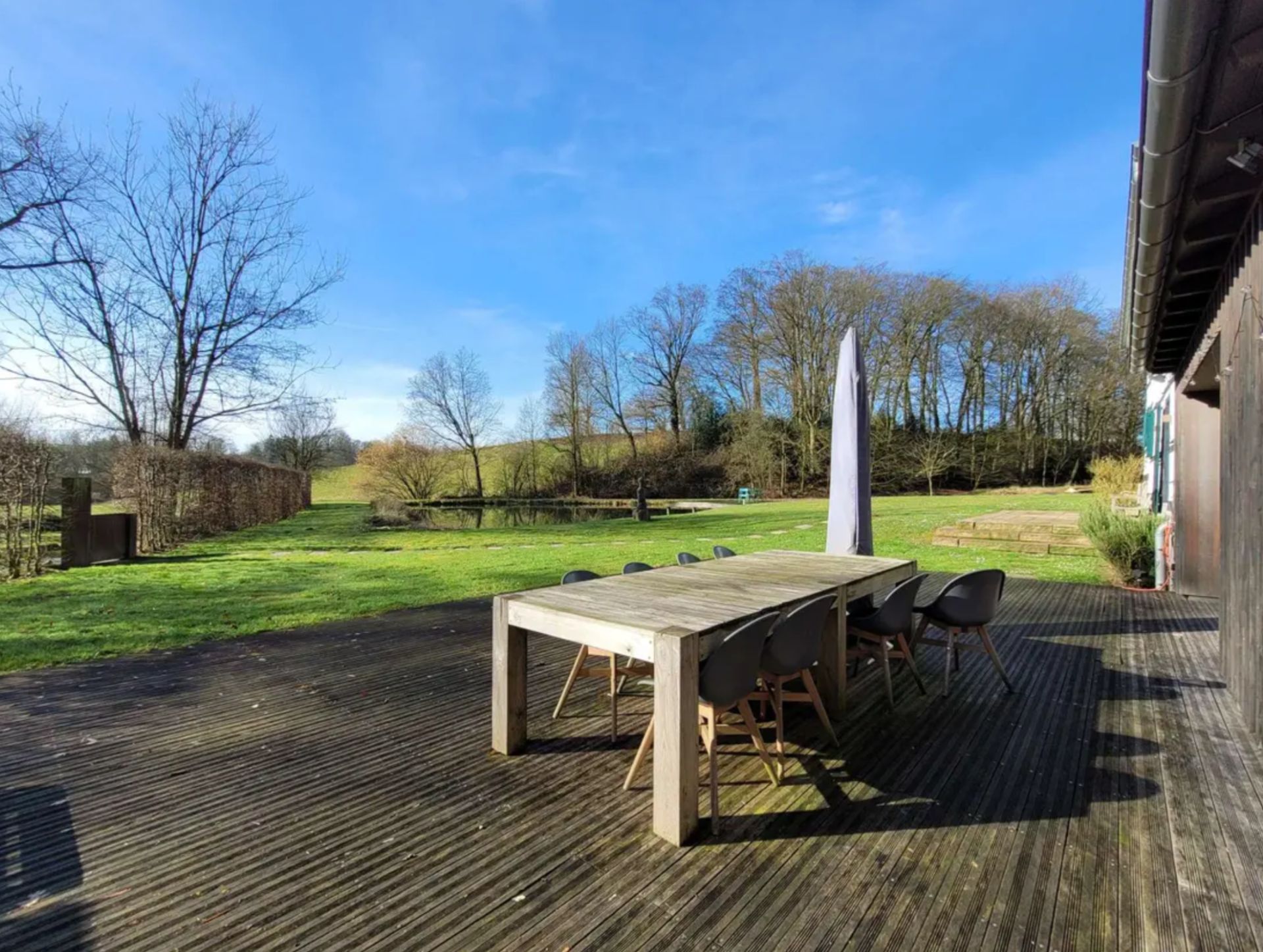 Holzterrasse mit Tisch und Stühlen, Blick auf Rasenfläche und Bäume im Hintergrund.