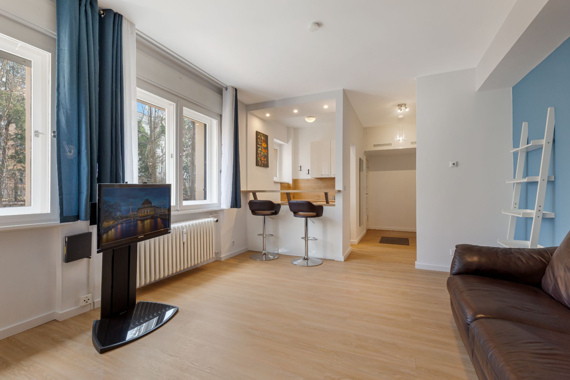 Helles Wohnzimmer mit braunem Sofa, Fernseher, Barhockern und offener Küche.