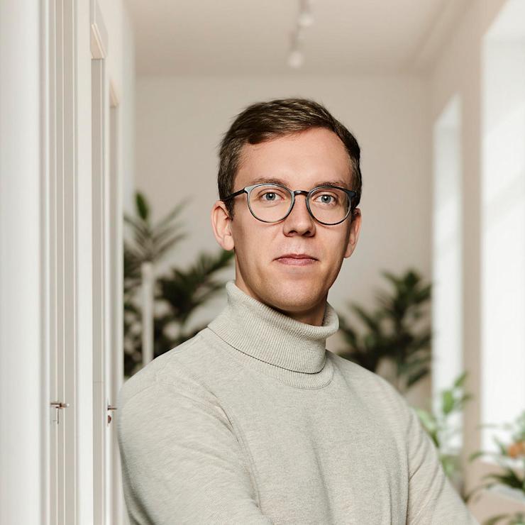 Mann mit Brille und Rollkragenpullover steht in einem hellen Flur mit Pflanzen.