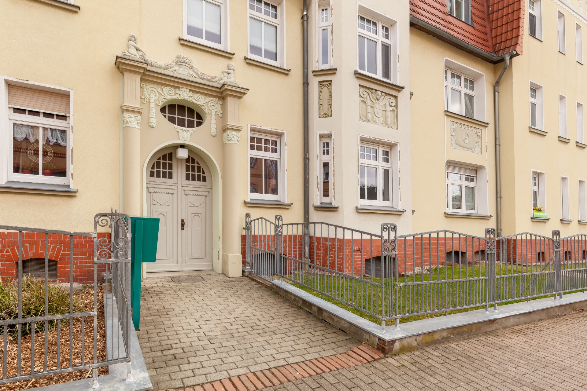 Eingang eines mehrstöckigen Wohnhauses mit Zaun, gepflastertem Weg und verzierten Fenstern.