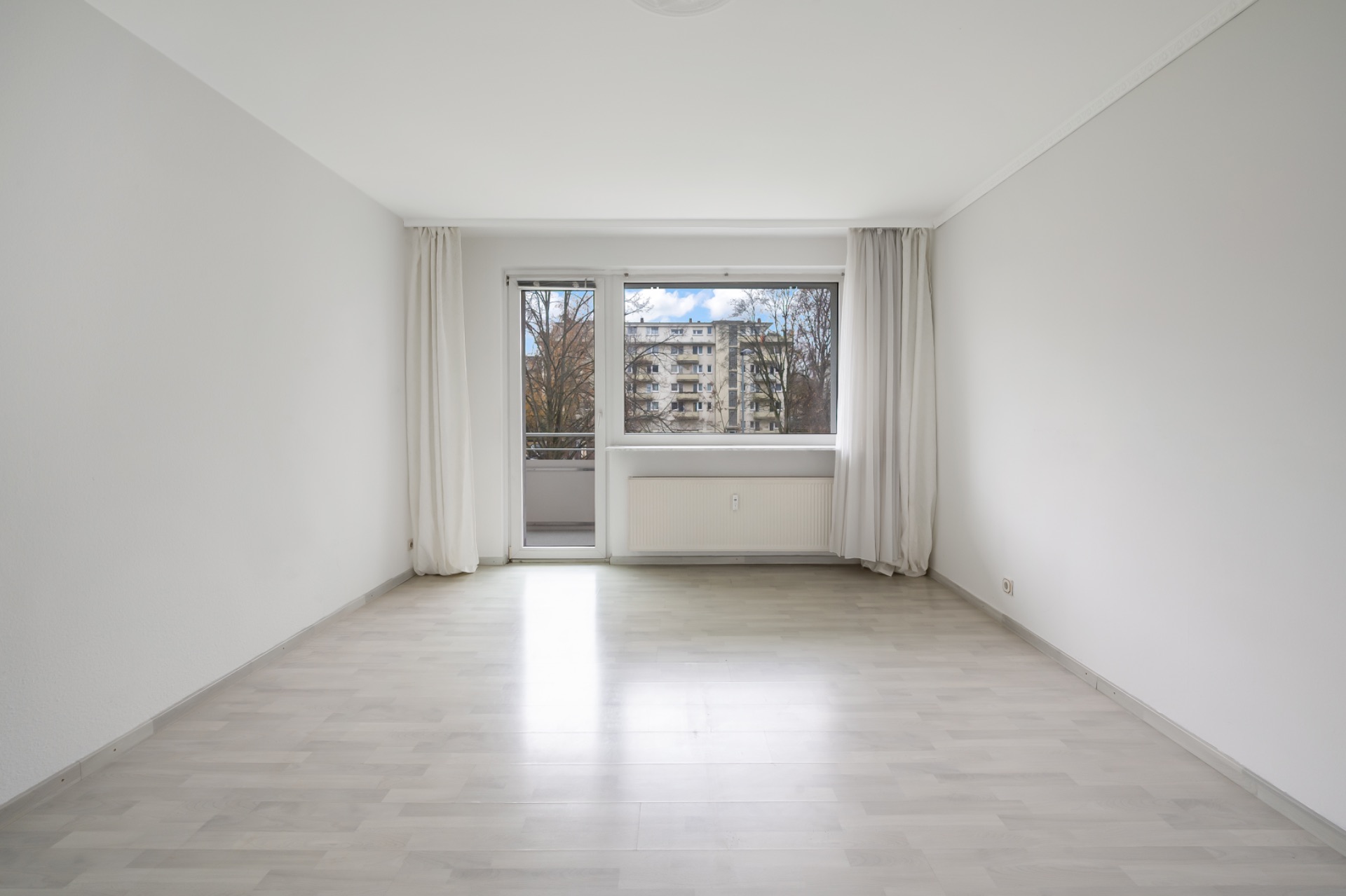 Helles, leeres Zimmer mit großem Fenster, Vorhängen und Blick auf Wohnhäuser.