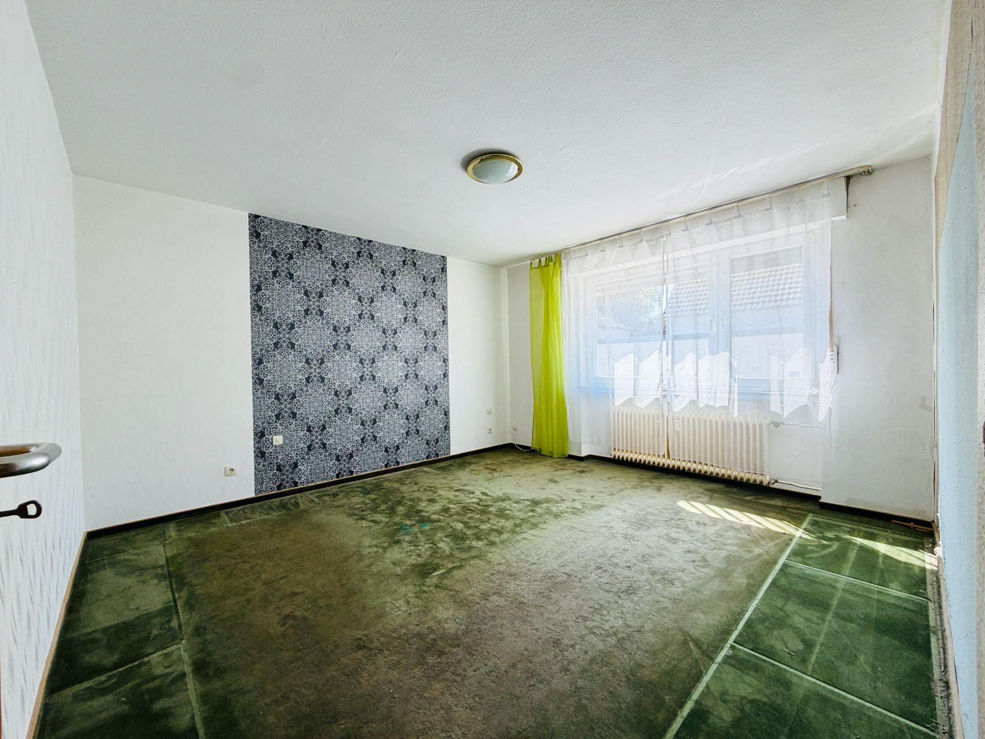 Leeres Zimmer mit grünem Teppich, gemusterter Wand und großem Fenster mit Vorhang.