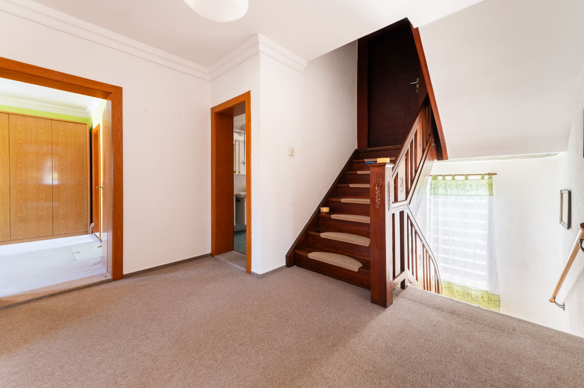 Flur mit Teppichboden, Holztreppe und Türen zu angrenzenden Zimmern, Fenster mit Gardine.