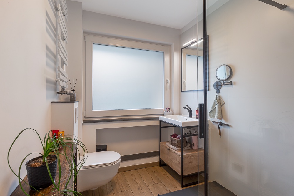 Modernes Badezimmer mit Toilette, Waschbecken, Glasdusche und großem Fenster.