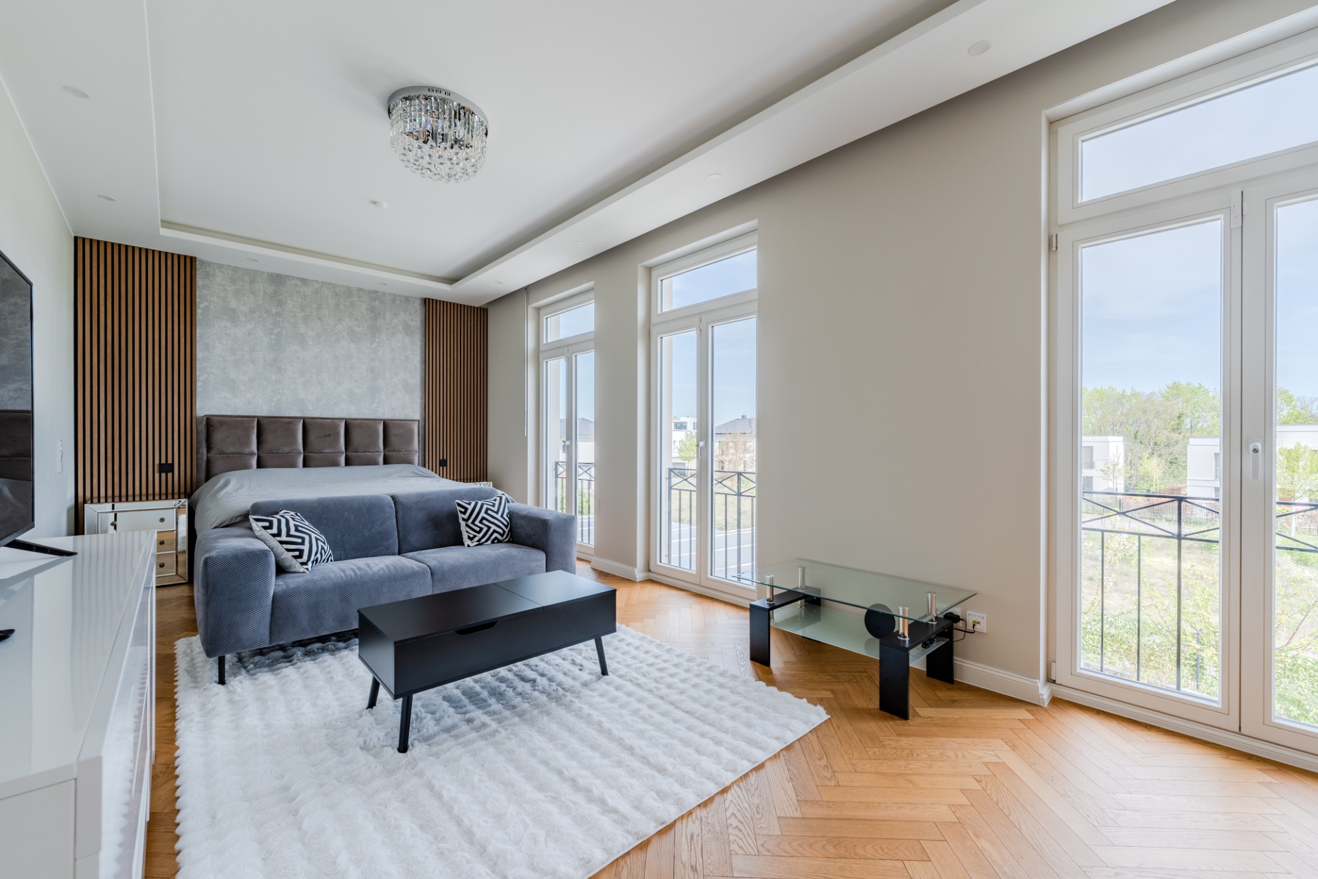 Modernes Schlafzimmer mit grauem Sofa, Doppelbett und großen Fenstern, die viel Tageslicht hereinlassen.