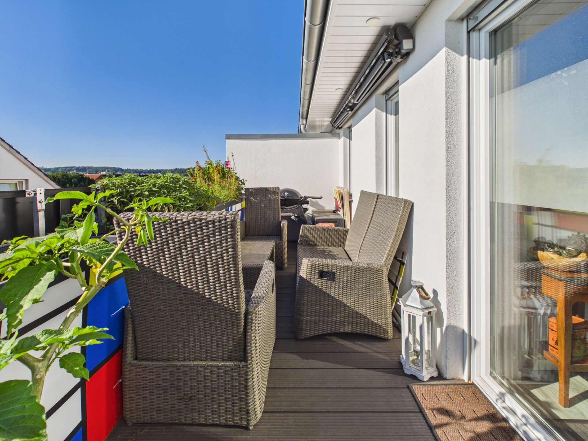 Balkon mit Rattanmöbeln, Pflanzen und einer weißen Wand, bei sonnigem Wetter.