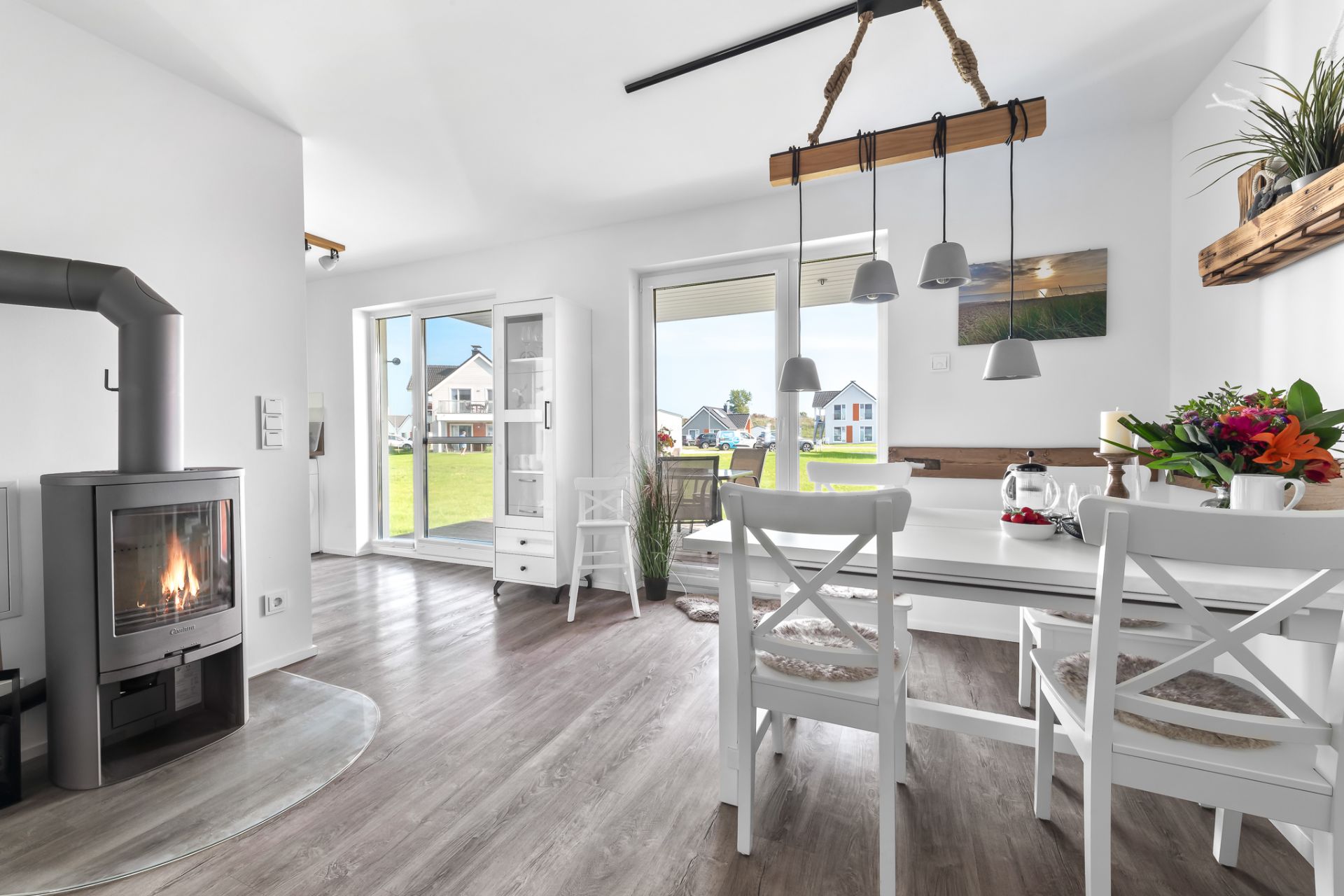Helles Wohnzimmer mit Kamin, Esstisch, weißen Stühlen und Zugang zur Terrasse mit Blick ins Grüne.