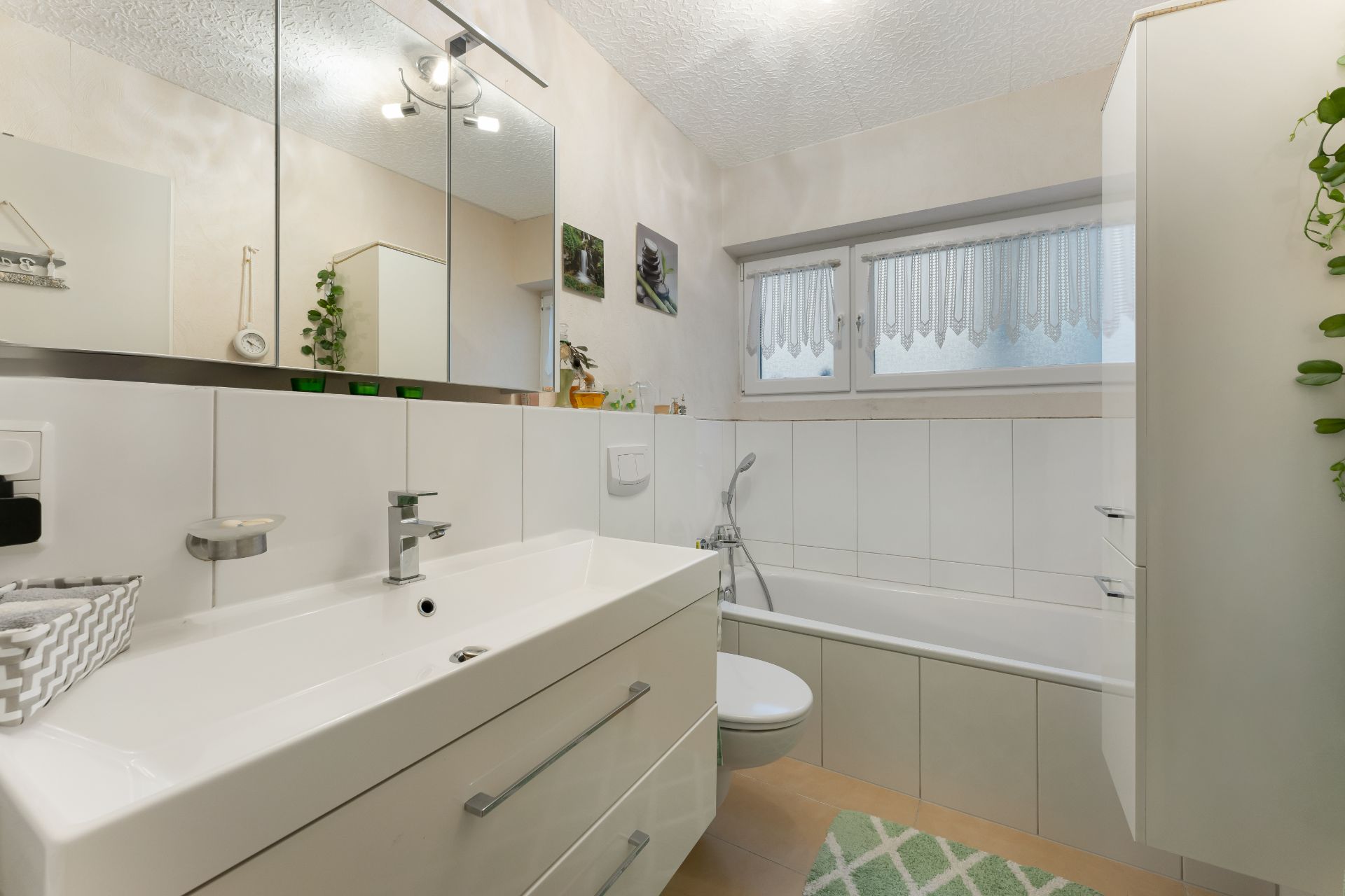 Helles Badezimmer mit Waschbecken, Badewanne, großem Spiegel und Fenster mit Gardine.