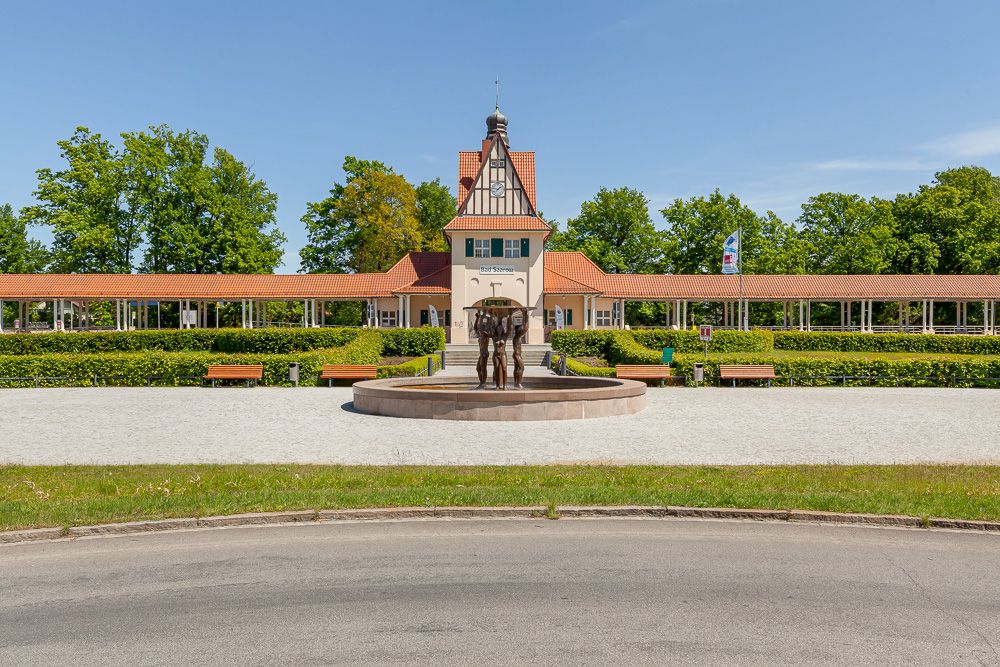 Gebäude mit rotem Dach, davor ein runder Brunnen und Bänke, umgeben von Bäumen.
