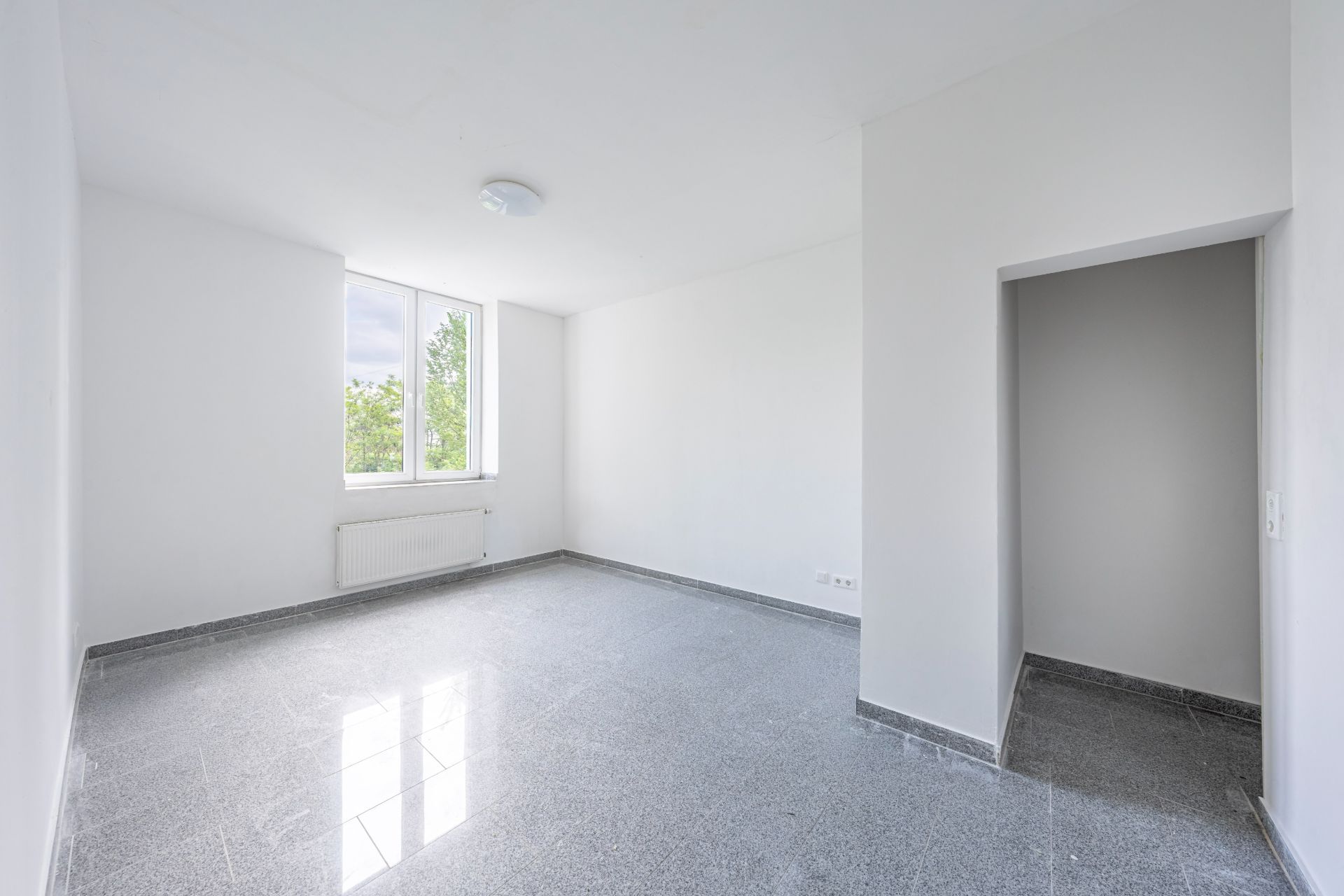 Heller, leerer Raum mit Fliesenboden, Fenster, Heizkörper und offenem Durchgang.