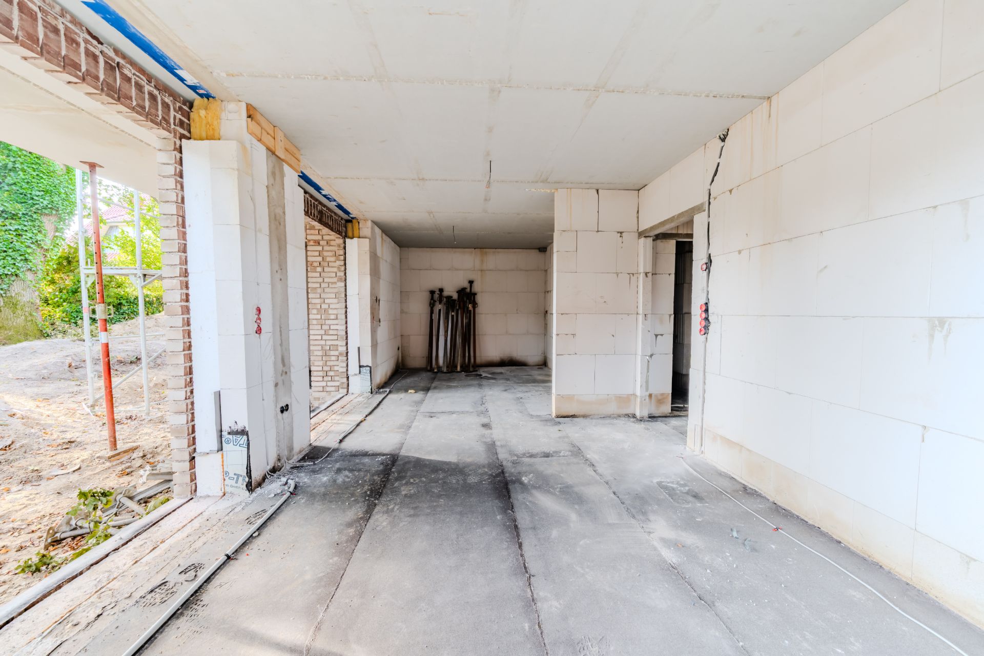 Rohbau eines Wohnraums mit weißen Wänden, offenem Boden und Baumaterialien im Hintergrund.