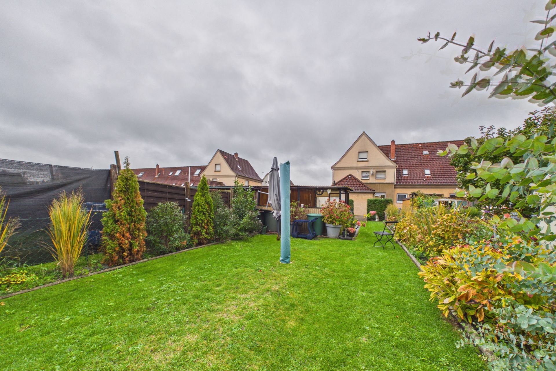 Grüner Garten mit Rasenfläche, Sträuchern, Gartenmöbeln und angrenzenden Häusern im Hintergrund.