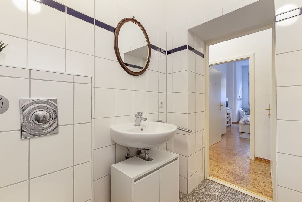 Heller, gefliester Waschraum mit rundem Spiegel, Waschbecken und Blick in den Flur.