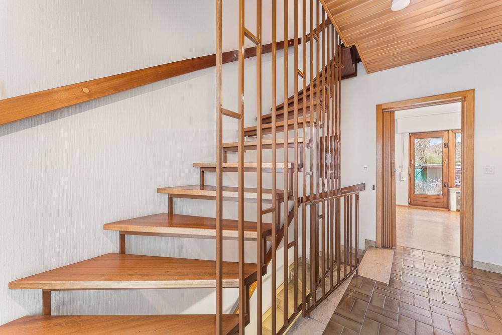 Holztreppe mit senkrechten Geländerstäben in einem Flur, Tür führt in helles Zimmer mit Balkon.